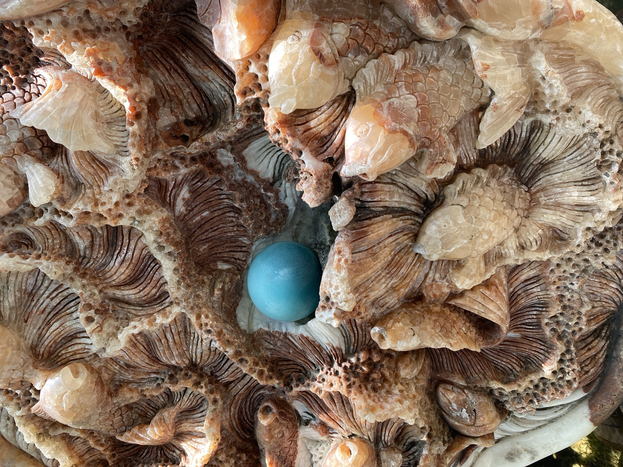 The image is a close-up of a carved stone relief sculpture depicting an underwater scene with fish and possibly coral or seaweed. A round, turquoise-colored stone or glass orb is prominently placed within a cavity of the sculpture. The sculpture itself is textured and appears to be made of a material with varying shades of brown, orange, and white, suggesting it might be onyx or alabaster. The lighting suggests it is outdoors, possibly in natural sunlight. The location in Đà Nẵng, Vietnam, is not identifiable from the image itself.