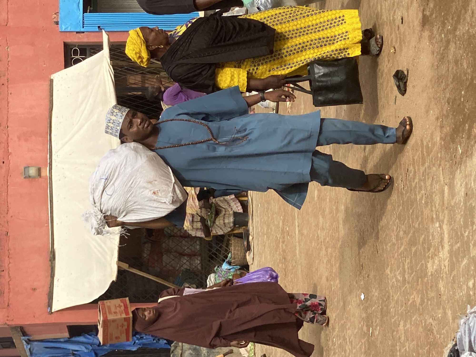 The scene depicts a street in Kafanchan B, Nigeria. A man wearing a blue robe and a patterned hat carries a large white sack over his shoulder. He wears brown sandals. A woman in a yellow patterned dress with a black wrap is standing behind him and has a black purse hanging off her arm. A woman in a brown robe walks in the opposite direction. The ground is dirt. Various objects and people are partially visible in the background. A building painted red is on the left. A white cloth is stretched above the man’s head, acting as a canopy. Blue sheeting and cardboard boxes are visible under it.