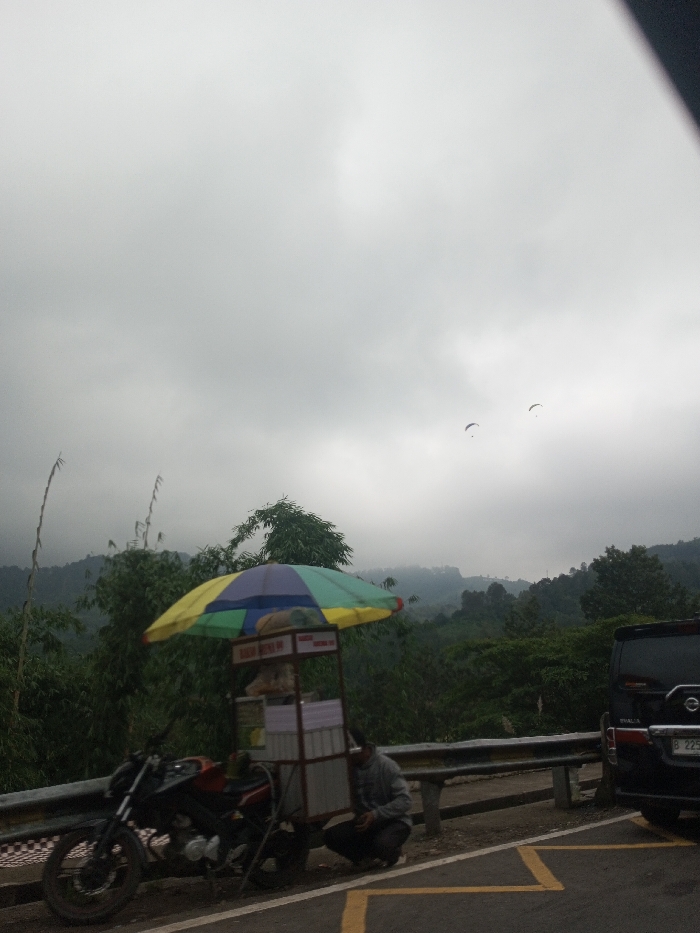 The image depicts a roadside scene located in Jabalpur, India, under an overcast sky with a mountainous, forested background. In the foreground, a dark-colored motorcycle is parked on the shoulder of an asphalt road, positioned to the left of a grey metal guardrail. Attached to the motorcycle is a mobile food cart, topped with a multi-colored umbrella (yellow, blue, green, red segments). The cart features a metal frame and white panels, displaying text that includes "BASO BOBA" and partially visible "BASO BA" on a lower sign. A male individual, wearing a grey hooded sweatshirt and dark pants, is squatting beside the food cart, facing the cart. To the right, a black SUV or van is parked, with its rear visible. The license plate on this vehicle begins with "B 225", and the text "THALIA" may be visible above it. Yellow road markings are present on the asphalt in the bottom right corner. In the mid-ground, dense green hills and trees rise, partially obscured by mist or haze. Two small paragliders are visible against the cloudy sky in the upper mid-ground.