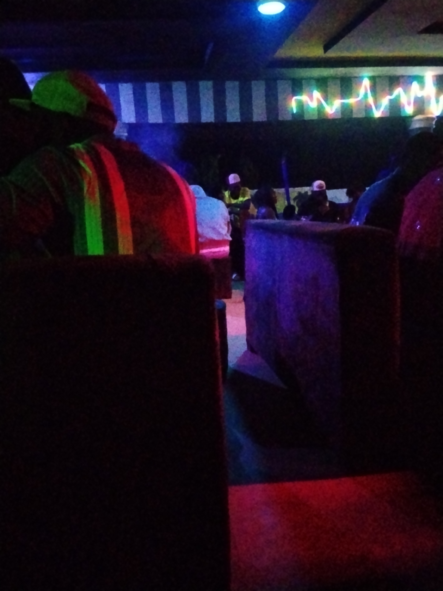 The image depicts an indoor scene at night, likely a club or entertainment venue in an unknown city. The lighting is dim and colorful, with neon lights and spotlights creating a vibrant atmosphere. In the foreground, a person wearing a cap and a striped shirt is visible, with green and red lights illuminating their back. In the background, several other people are seen in silhouette, some wearing caps and others sitting. The environment features dark furniture, including what appears to be a large, upholstered seating area with purple lighting. A wall with vertical stripes and a colorful light display resembling a sound wave is also present. The overall impression is that of a lively social gathering in a dimly lit, modern space.