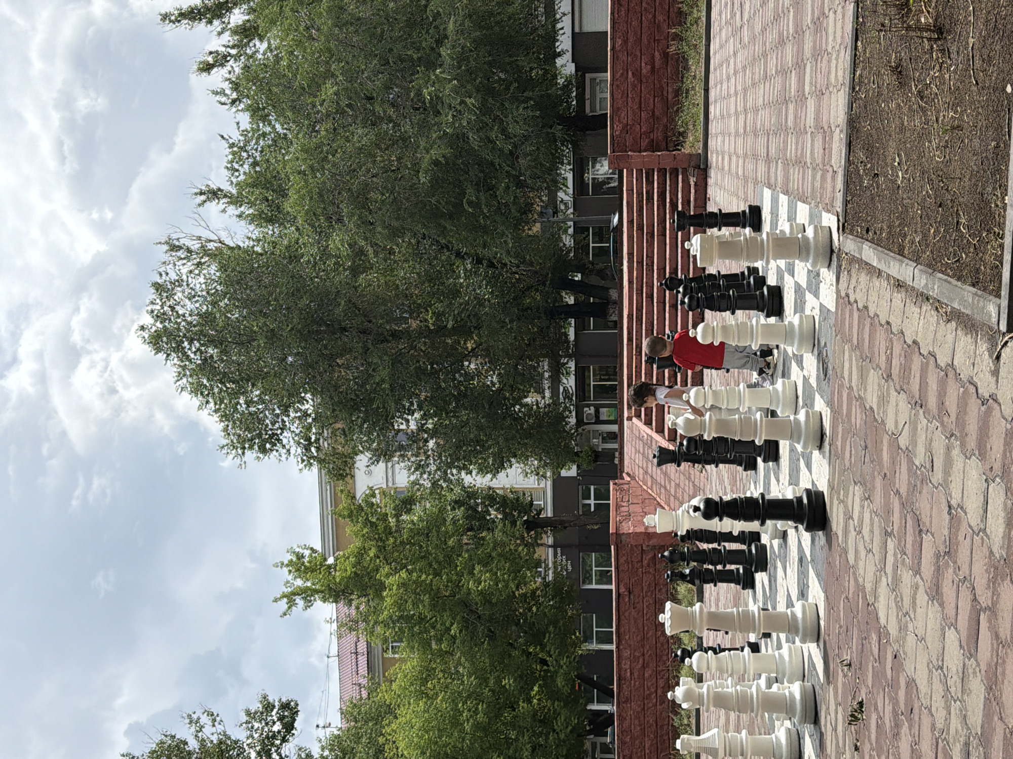 In Karaganda, Kazakhstan, two young children are observed playing a large outdoor chess game. One child, wearing a red shirt and grey pants, stands on the chessboard, positioned to the right of the frame, looking towards the pieces. The other child, partially visible, stands behind them on the chessboard, on the left side of the frame. They are surrounded by oversized black and white chess pieces, including pawns, knights, rooks, and queens. The chessboard is set on a patterned brick pavement. In the background, a multi-story building with a red-tiled roof and light-colored facade is visible behind a dense line of green trees. A red brick wall with steps leading up to it forms the boundary of the chessboard area. The sky above is overcast with a mix of white and grey clouds.