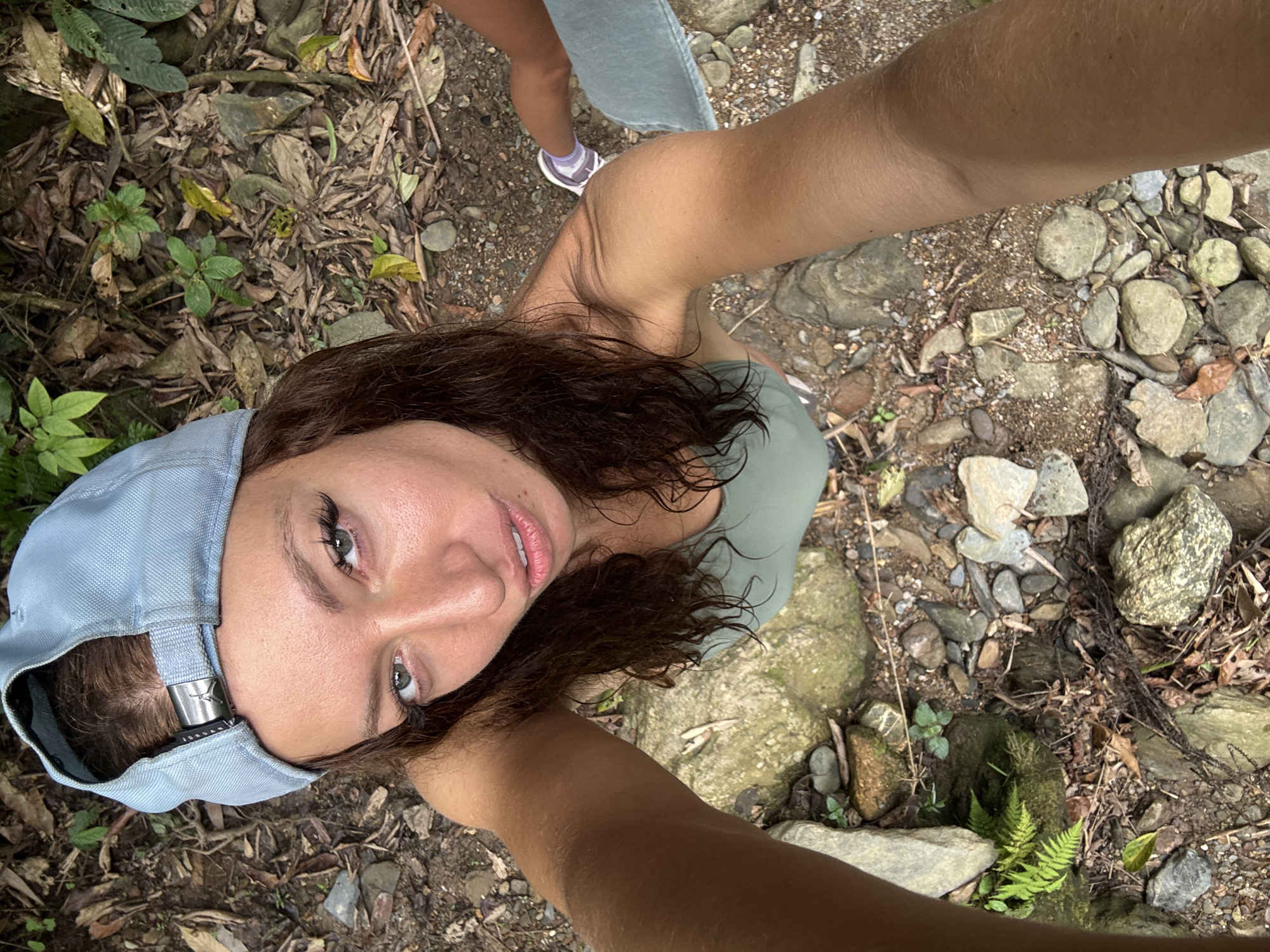 The image shows a first-person perspective selfie taken by a woman in a natural environment. The foreground features the woman's face wearing a light blue baseball cap, and she appears to be looking directly into the camera. Her arms extend towards the camera, framing the shot. In the midground, the background shows a rocky, foliage-covered area, indicative of a natural setting. A portion of another person's leg wearing jeans and a sneaker is partially visible in the top left corner. The ground is covered with rocks, leaves, and small plants. Based on the location context, it is possible that the photo was taken in Suối Giàng, Vietnam.