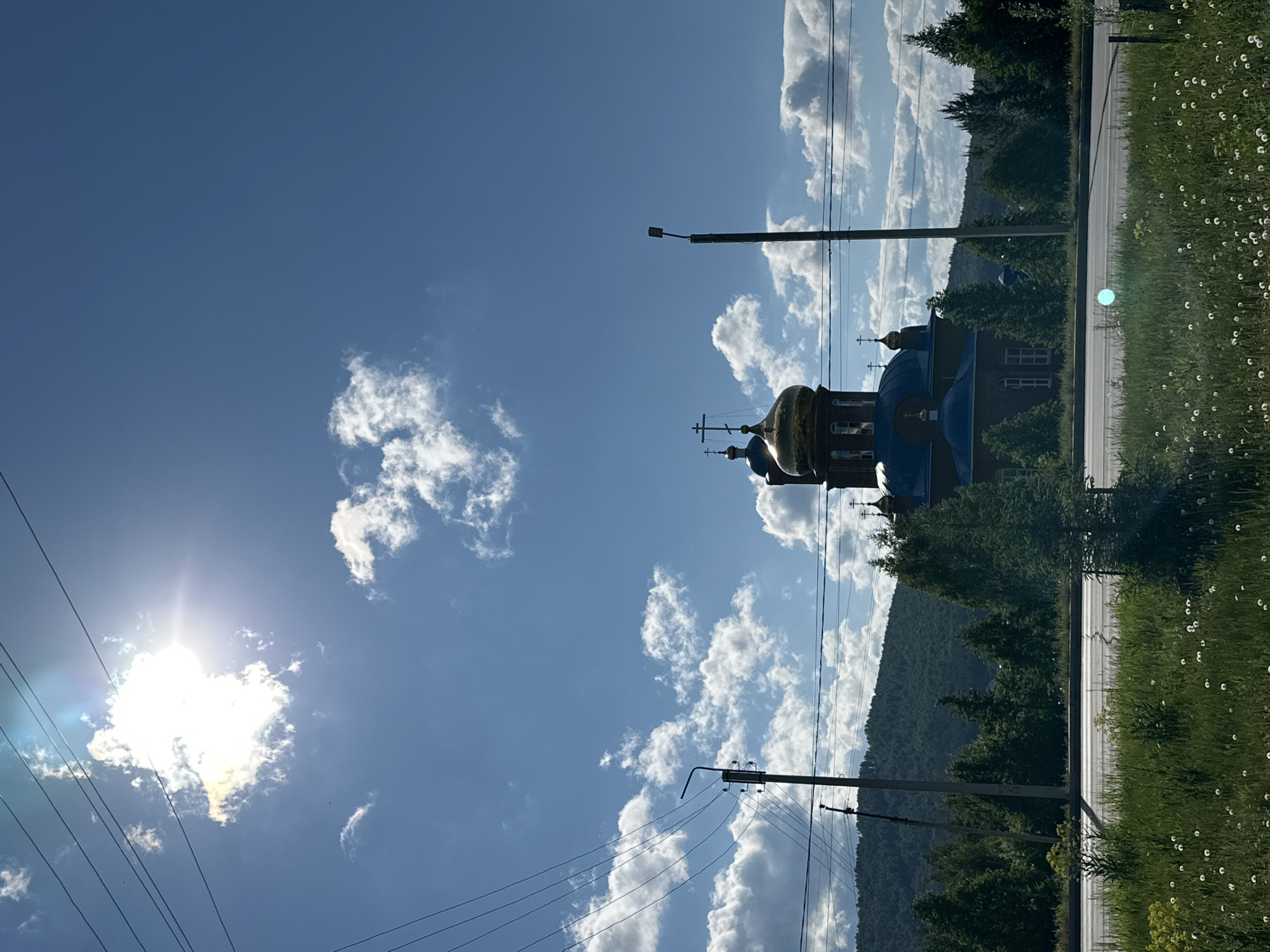 A daylight shot presents a landscape with a blue sky filled with white clouds, taken at an angle. A building with blue roofs and gold domes stands amongst green trees, with a hill rising behind them. Power lines are visible crossing the sky. The right side of the image shows a road, overgrown with green grass and small white flowers. The sun is positioned in the lower left corner, partially obscured by clouds. The location is Unknown, Unknown.