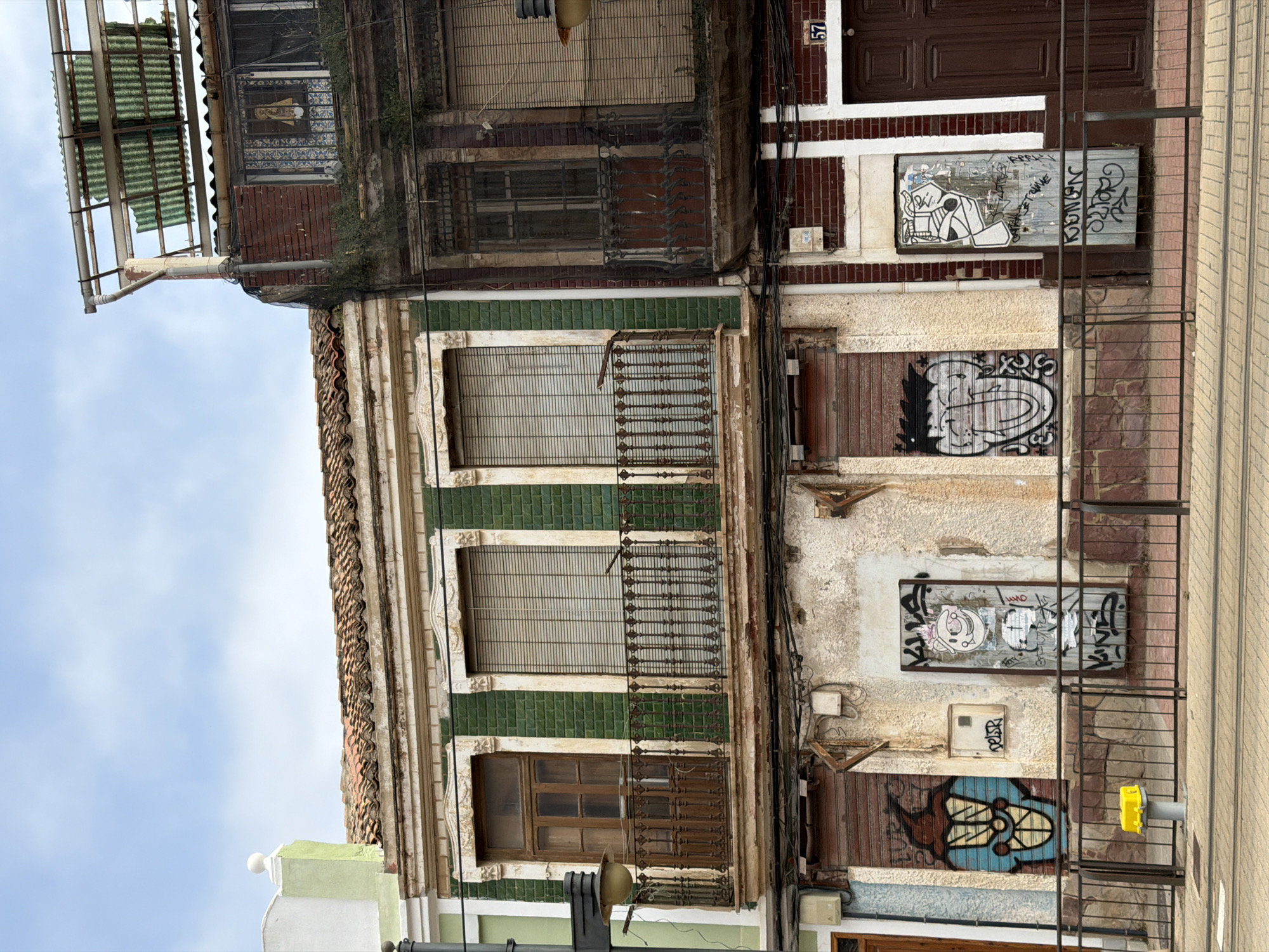 The image shows a rundown building facade in an urban environment. The building has multiple stories and is covered in graffiti. On the ground floor, there are roll-up doors, one of which is covered in a blue and black cartoon-like graffiti, and another that appears to be a metal door with various tags and a drawing of a pig-like character. Above, there are balconies with ornate railings. The windows are covered with blinds. The building appears to be old and in a state of disrepair. The sky is visible in the background, with some clouds. The overall impression is one of urban decay and neglect.