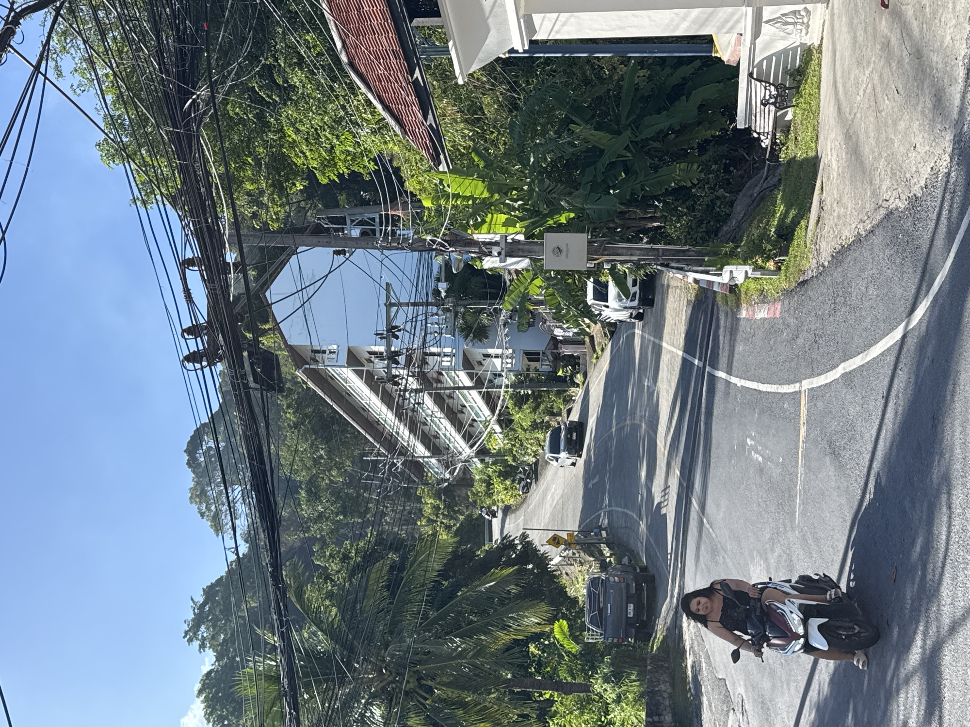 A woman rides a scooter on a paved road in Patong, Thailand. The road curves uphill, flanked by lush green foliage and buildings. Several cars and motorcycles are parked or driving on the road. Overhead, a complex network of power lines crisscrosses the sky. A white building with multiple balconies and white railings is visible on the right side of the road, along with a red-tiled roof. The woman is wearing a black sleeveless dress and is looking forward. She is wearing white sandals. Sunlight casts shadows on the road.