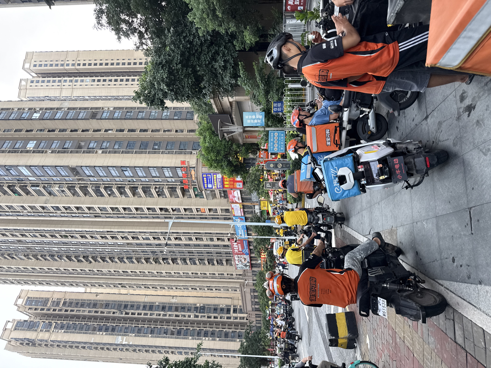 The image captures a street scene in Haizhu District, China, dominated by delivery workers and their electric scooters parked along the sidewalk. Tall, modern buildings with numerous windows line the background, suggesting an urban environment. The delivery workers, most wearing orange or yellow vests and helmets, are gathered near their vehicles. Some appear to be resting, while others are interacting.
The scooters are equipped with large insulated boxes, a common sight for food and package delivery services. Visible text on some of these boxes includes "淘宝闪购" (Taobao Flash Purchase) and "小牛电动" (Niu Electric), indicating the brands or services involved. Other signage in the background includes street names and advertisements.
The time of day appears to be daytime, with ample natural light, though the sky is overcast, creating a diffused lighting effect. The overall atmosphere seems to be one of organized waiting, typical of delivery hubs or staging areas where couriers might congregate between assignments. The presence of many individuals and their vehicles suggests a busy period for delivery services in this densely populated urban area.