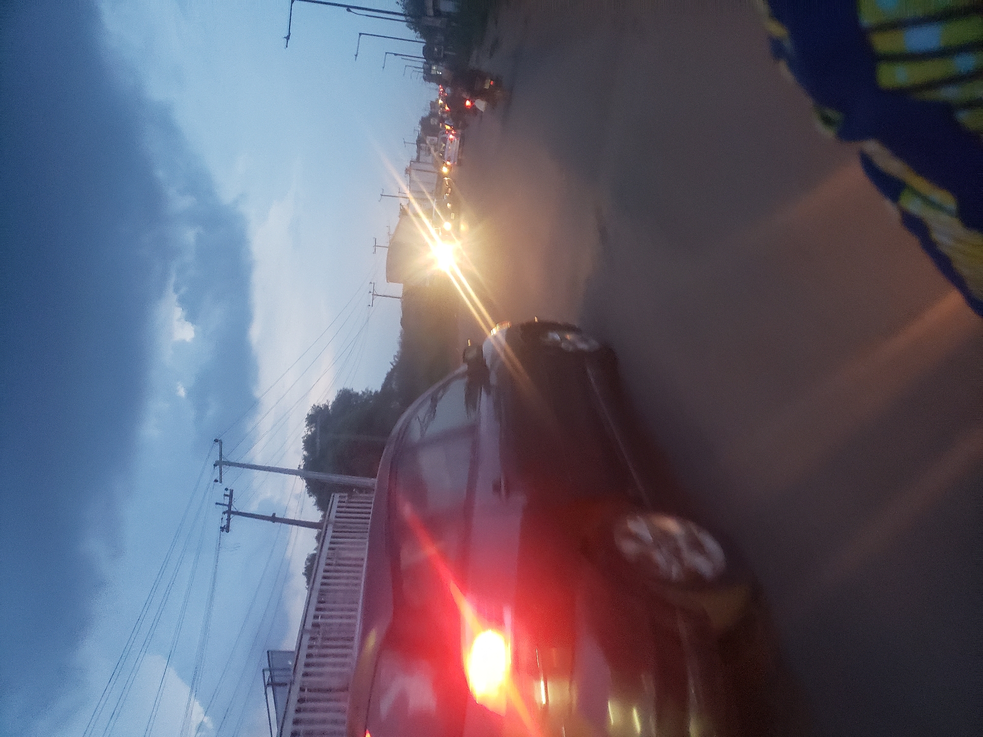 The image depicts a road scene in Ida, Nigeria, at dusk or dawn. The sky is a gradient of blue, with dark clouds on the left side of the frame. Power lines and utility poles are visible against the sky. A road stretches into the distance, with vehicles present on it. Bright headlights suggest traffic moving towards the camera's position. A red car is partially in the frame, occupying the lower central part of the image. The edge of a building or structure is present on the left side, alongside the road. A partially visible subject is also present on the upper right, with colorful patterns on fabric.