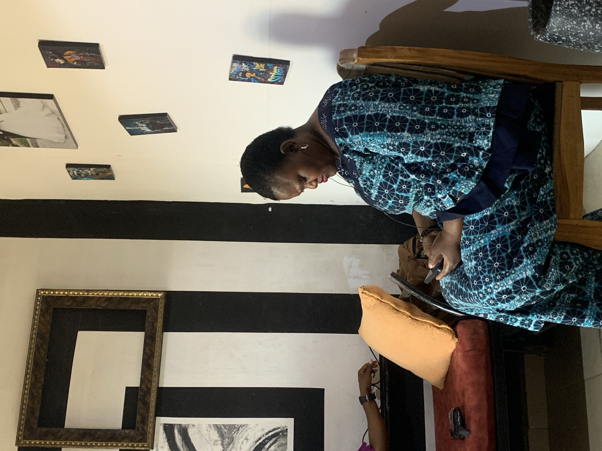 A woman with dark hair, fair skin, and wearing a blue and white patterned dress and earbud headphones sits on a wooden chair indoors. She is looking down at a black smartphone she holds in her hands.
Behind her is a white wall with several framed photos hanging on it. The wall transitions into a black and white striped pattern near the bottom. A large empty gold frame is also affixed to this lower section of the wall. A small section of an abstract artwork is visible below the frame.
To the right of the chair, an orange pillow rests on a red cushion. An arm holding a pair of glasses extends upwards from the bottom of the frame.
The location is Gura Top, Nigeria.