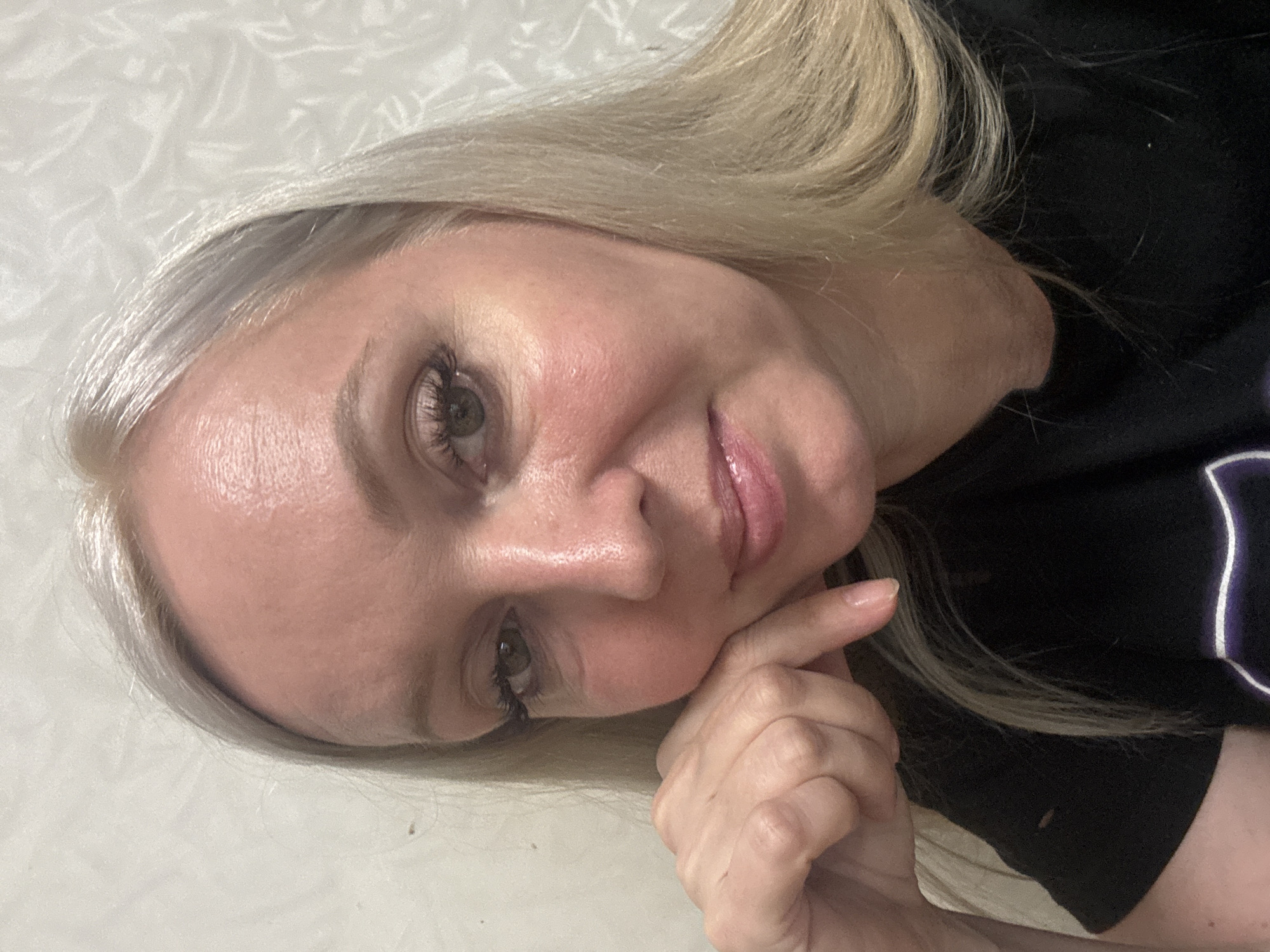 A close-up, waist-up selfie shows a fair-skinned woman with blonde hair, resting her chin on her right hand. She is wearing a black t-shirt. The background is a textured, light-colored wall. The image does not contain any information that would allow for the determination of its location in Komsomolsk-on-Amur, Russia.