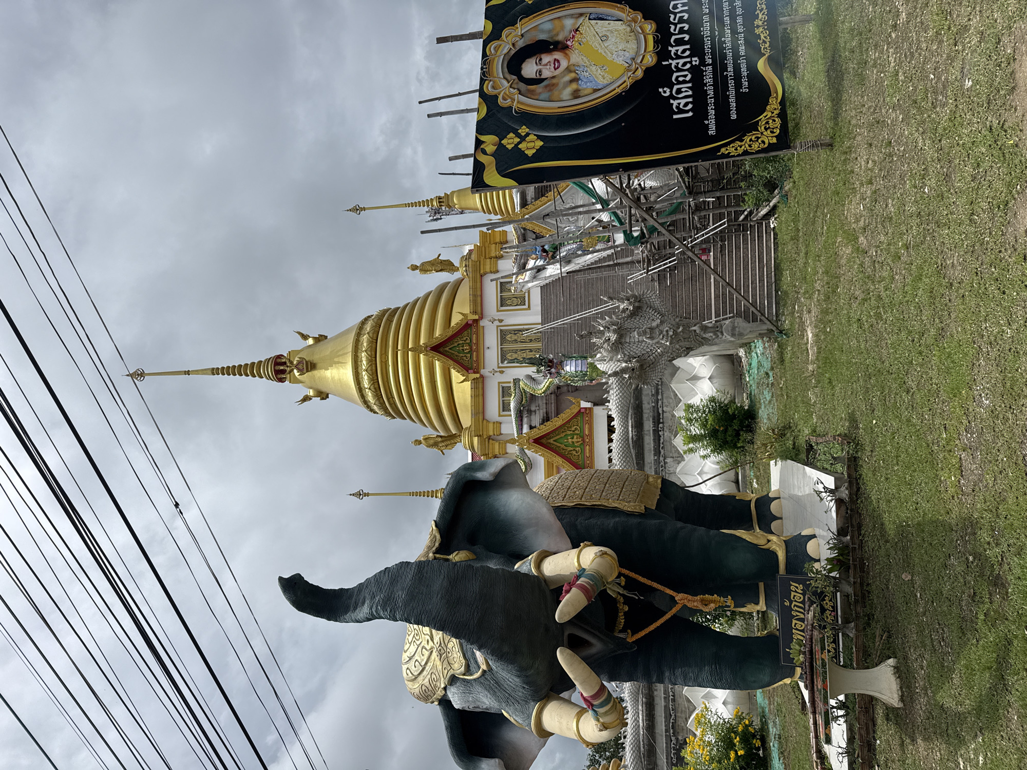 In Ban Sida Ngam, Thailand, a large, dark green elephant statue with gold accents stands prominently on a grassy lawn in front of a Buddhist temple. The elephant has its trunk raised, adorned with gold armbands on its legs and tusks. Behind the elephant, a golden tiered stupa with a spire dominates the temple structure, which features white walls, ornate roofs, and a staircase lined with dragon sculptures. To the right of the temple entrance, a black banner displays a portrait of a woman in traditional Thai attire, accompanied by Thai script. The sky above is overcast with gray clouds, and power lines are visible in the upper portion of the image. A small white table and planter are placed in front of the elephant statue. The scene suggests a cultural or religious site, possibly commemorating a significant figure.