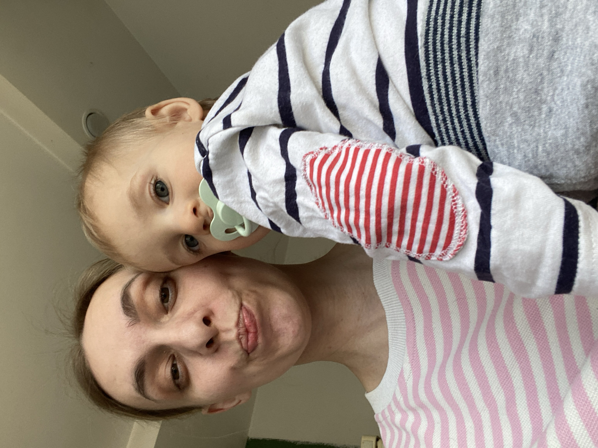The image displays an adult female holding an infant in a close-up, indoor setting within the city of Chaikovsky, Russia. The female, positioned on the left, has light brown hair and is wearing a white long-sleeved top with horizontal pink stripes. Her lips are pursed in a puckering expression, and her gaze is directed forward. The infant, positioned on the right and held by the female, has blonde hair and blue eyes, with a light green pacifier in its mouth. The infant is wearing a white long-sleeved top featuring dark blue horizontal stripes and a red and white striped elbow patch on the visible left sleeve. The background is a light-colored ceiling, with a circular fixture partially visible in the upper right quadrant. A faint green vertical element is discernible on the far left edge of the frame.