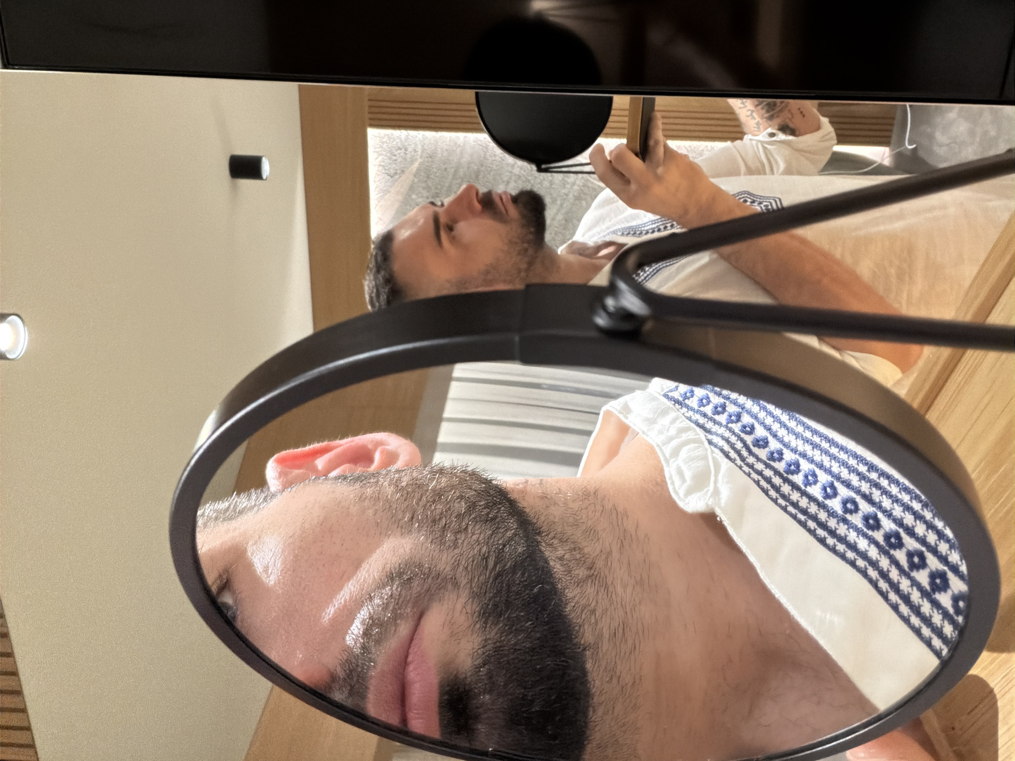The image depicts a man reflected in a round mirror. The upper part of the frame includes a view of the man looking at a handheld device, presumably a smartphone. The background includes what appears to be a room interior with wood-paneled elements. The man is wearing a white garment with blue patterned trim. The mirror frame is black and has supporting arms. The location in Baldevo, Bulgaria is not discernible from the visual information provided.