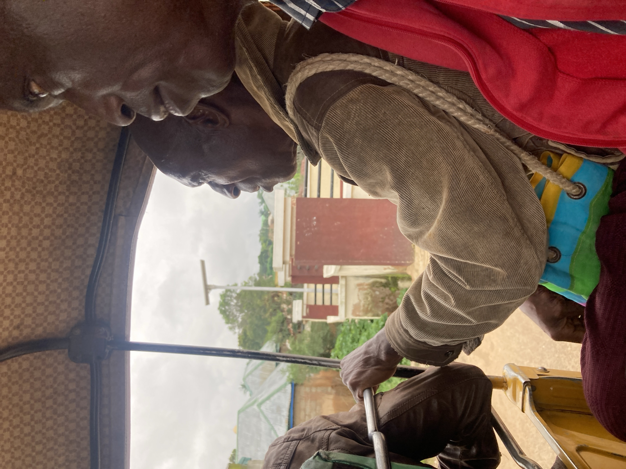 The image shows two Black men inside a motorized rickshaw, likely a Bajaj or similar model, in Jos, Nigeria. The driver, an elderly man with a dark complexion, is wearing a light brown corduroy jacket over a colorful, possibly fabric bag strapped to his shoulder. His right hand grips a metal bar, presumably part of the steering mechanism. The passenger, also a Black man with dark skin, is partially visible to the right, with his head and shoulder in the foreground. He is wearing a dark blue and white striped shirt with a red jacket over it. The rickshaw's interior ceiling has a checkered pattern. The exterior view through the open side of the rickshaw reveals buildings with corrugated metal roofs, green foliage, and what appears to be a street lamp in the background. The sky is overcast, suggesting cloudy weather. The overall scene depicts everyday transportation in an urban environment.