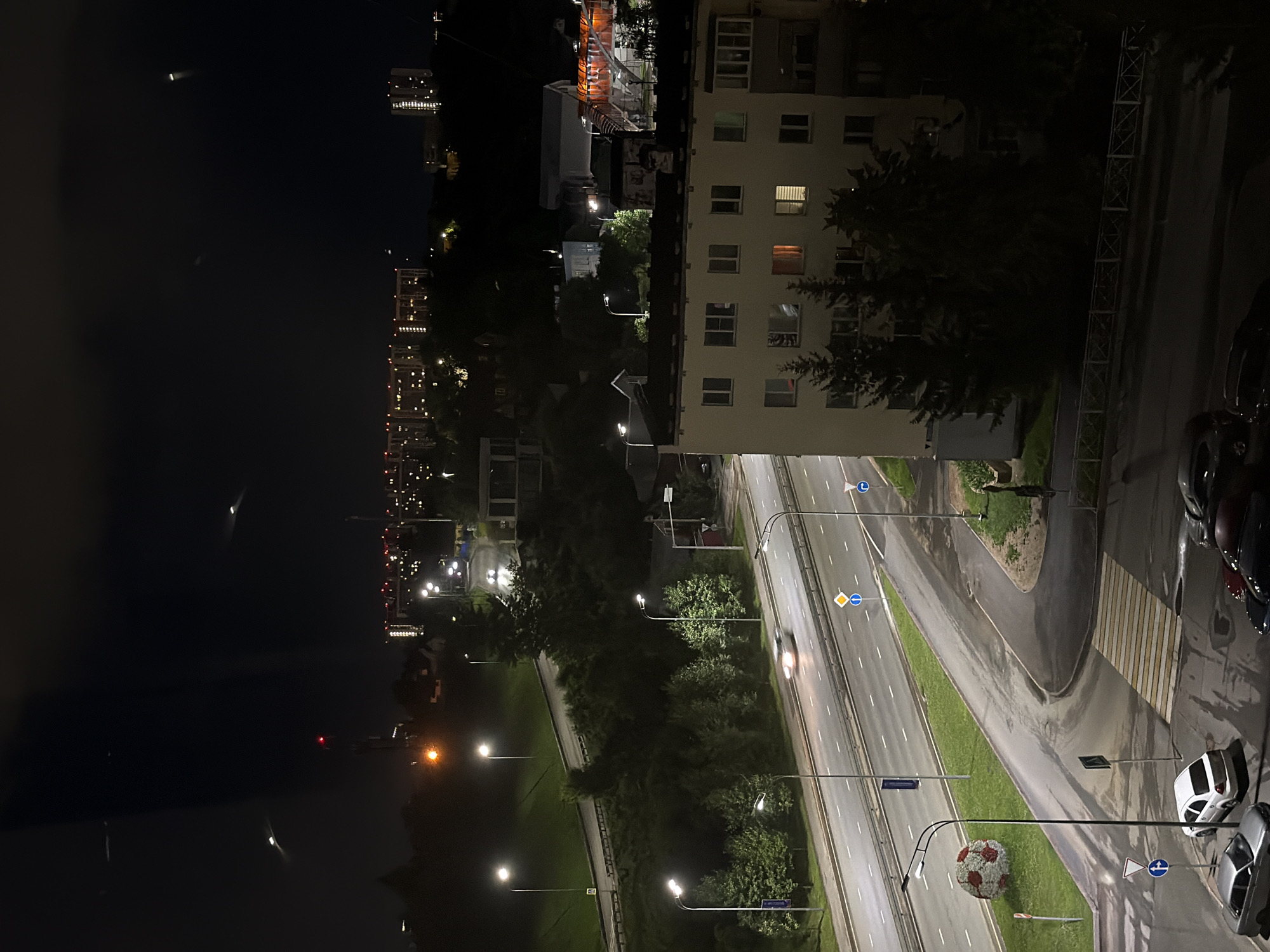 The image depicts a nighttime urban scene in Kazan, Russia, likely from a high vantage point. A multi-lane road with streetlights runs through the center, with a few cars visible on it, one appearing to be in motion. Adjacent to the road is a grassy embankment and a line of trees. A distinctive large spherical sculpture resembling a soccer ball, covered in red and white patterns, is situated on the grass. In the foreground, a residential building with illuminated windows stands, and several parked cars are visible on the asphalt below. In the background, more city buildings with illuminated windows and lights are spread across the dark, hilly landscape, hinting at the broader urban sprawl of Kazan. The sky is dark with some faint lights or atmospheric phenomena, possibly streaks from passing aircraft or distant flares.