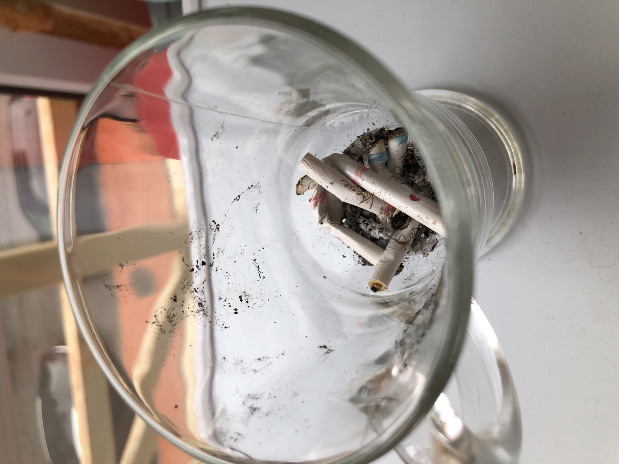 An indoor close-up, high-angle shot shows a glass ashtray filled with several cigarette butts. The ashtray is clear glass, and the cigarette butts are partially burnt, with ash and debris accumulated within the glass. Some of the cigarette papers have colorful designs, including pink and blue accents, and text that appears to read "Love." The background is blurred, suggesting a shallow depth of field, and shows what appears to be a window with wooden blinds and a reddish-orange wall. The scene is likely in Saint Petersburg, Russia, given the context provided.