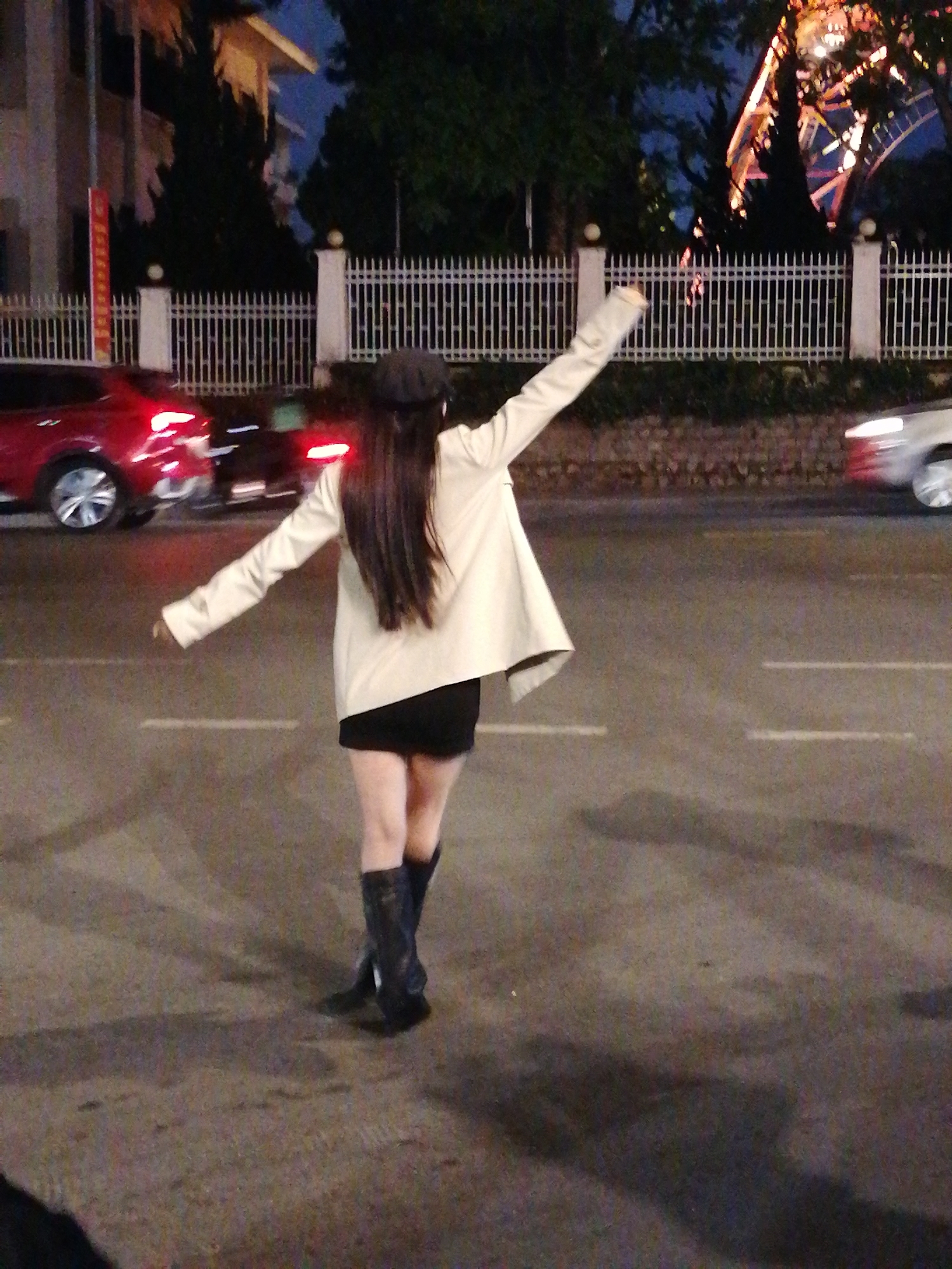 An individual wearing a black beret, a cream-colored jacket, a black skirt, and black boots stands on a paved road in Da Lat, Vietnam. The person's back is to the camera, and their arms are outstretched, with the right arm pointing upwards and to the left, and the left arm extended outwards and to the left. The person's hair is long and dark. The background features a white fence, trees, and a building. In the distance, a Ferris wheel-like structure is illuminated. Red and white traffic lights are visible, along with moving vehicles, suggesting a nighttime setting on a city street.