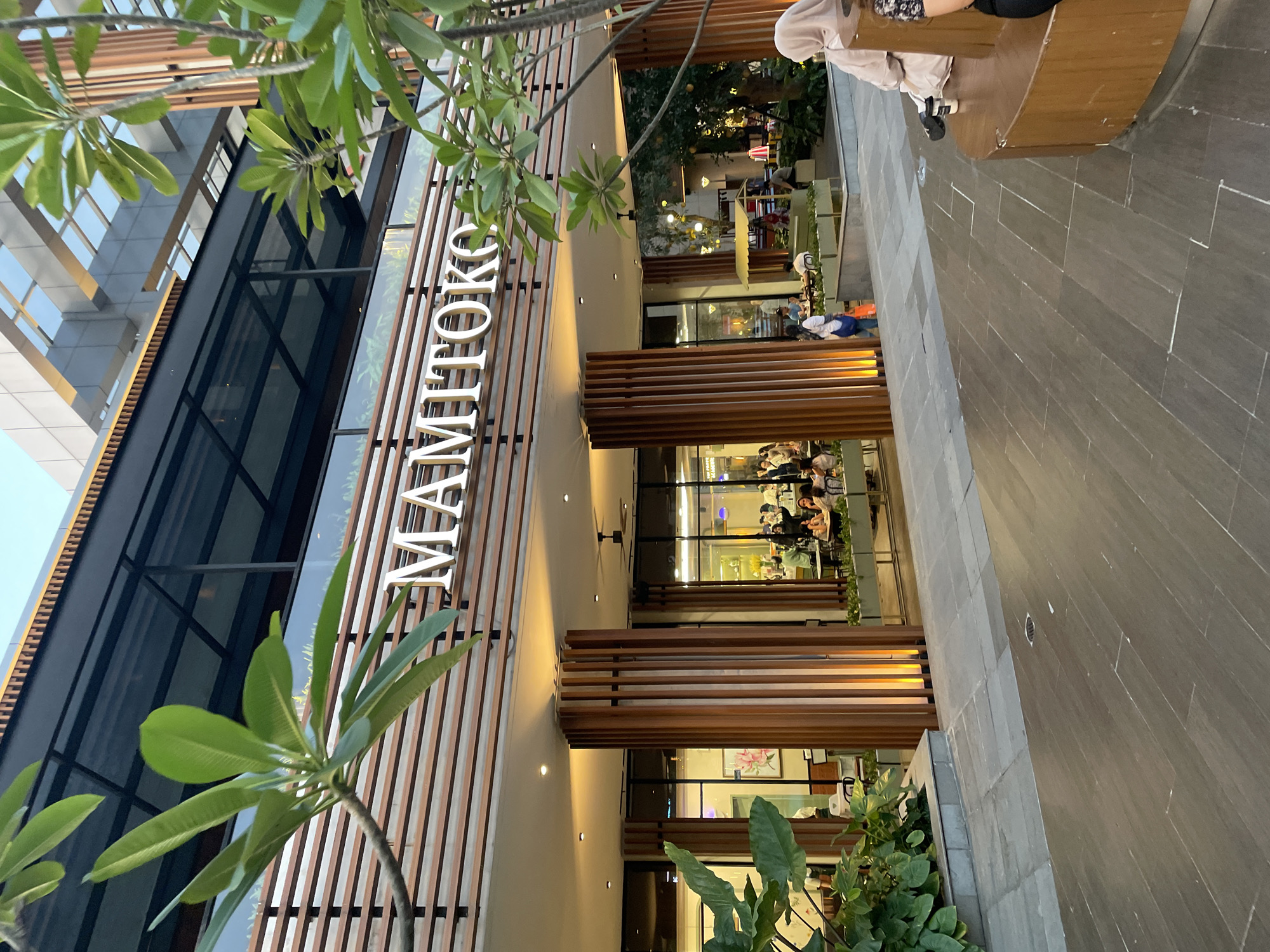 The image shows the exterior of a restaurant called "MAMITOKO". It is located in North Jakarta, Indonesia. The restaurant has a modern design with wooden accents and large glass windows. People are seated at tables inside the restaurant. The restaurant's name is displayed vertically in large white letters on the exterior. The scene is outdoors, with pavement visible in the foreground and foliage on either side of the restaurant entrance. Part of a bench and a person's leg are visible on the upper right edge of the image.