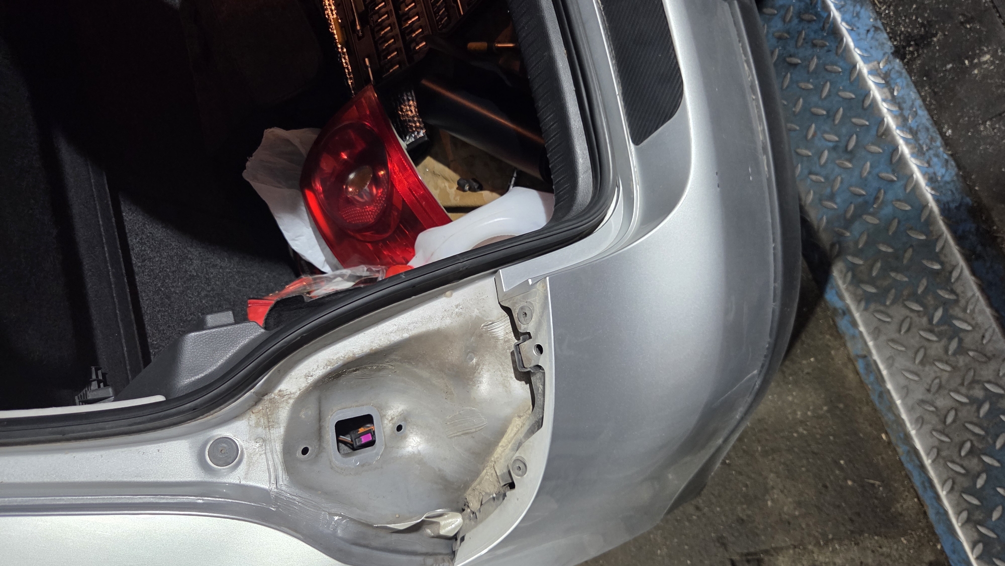 An interior shot of an open car trunk shows a red tail light and a white plastic container on the right side. The trunk's interior is lined with dark fabric. On the left side of the image, the exterior of the car is visible, showing a silver-painted quarter panel with a detached tail light assembly. Underneath the car, a textured metal surface with blue accents is visible.