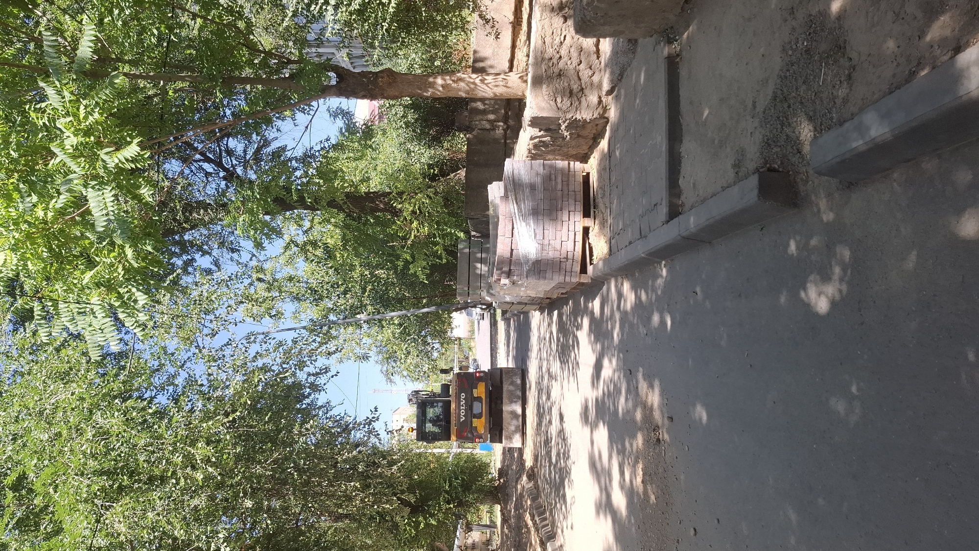 The image depicts a street scene in Shymkent, Kazakhstan, with a yellow Volvo excavator parked on the asphalt. The excavator is positioned on the left side of the road, facing away from the camera. To the right of the excavator, there are stacks of paving stones and concrete blocks, suggesting ongoing construction or repair work. The area is lined with trees, casting dappled shadows on the ground, and a sidewalk is visible along the right side of the road. In the background, there are buildings and a few parked cars, indicating an urban environment. The sky is clear and blue, and the overall lighting suggests it is a sunny day. The presence of construction materials and machinery points to infrastructure development or maintenance in the city.