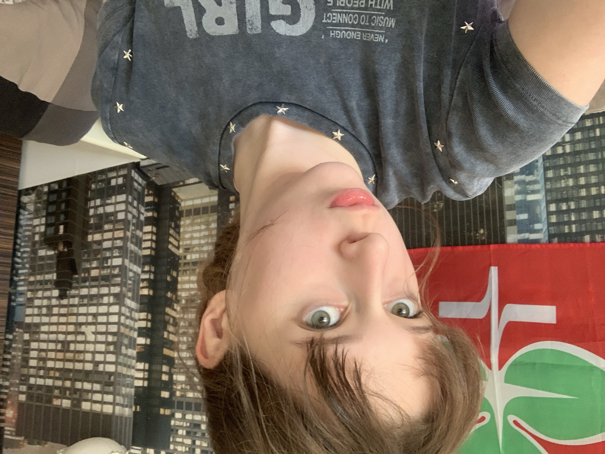 A woman with light skin, green eyes, and brown hair is seen in a selfie-style photograph. She is wearing a gray t-shirt with the word "GIRL" printed on it, along with the phrase "NEVER ENOUGH, MUSIC TO CONNECT WITH PEOPLE". The t-shirt is adorned with small, star-shaped embellishments around the collar.
Behind her is a black-and-white cityscape wallpaper, and a red flag with a stylized white cross and green leaf design.
There is no information about the location being Vozdovac Urban Municipality, Serbia in the image.