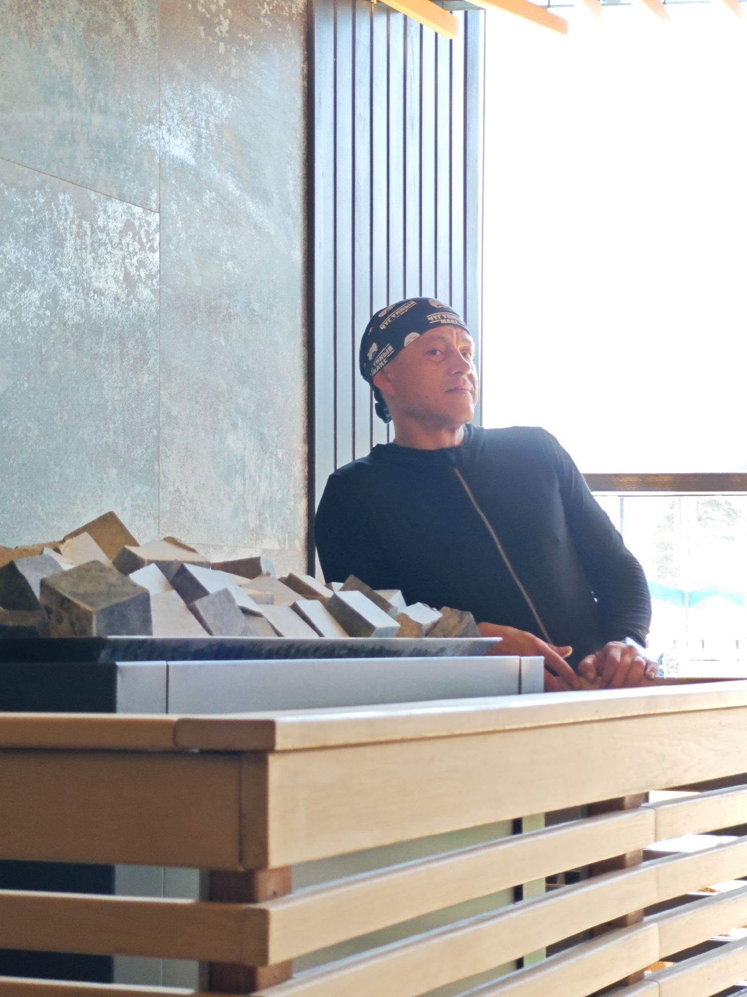 A middle-aged man with a bald head, wearing a black long-sleeved shirt and a black bandana with white writing, is seated behind a counter. The counter has a display of small, rectangular stones of varying colors and textures. The counter itself is made of light-colored wood with horizontal slats. Behind the man is a wall with alternating panels of textured, gray material and vertical wood slats. The background is brightly lit, obscuring details of the view outside the window. The location appears to be indoors. There is no specific information available to confirm it is in Votkinsk, Russia.