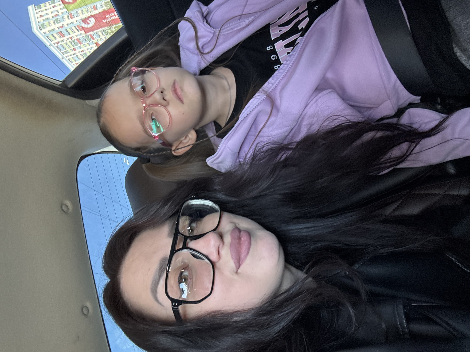 The image features two females in a vehicle. The woman in the foreground has dark hair and is wearing glasses and a black leather jacket. The girl in the background has her hair braided and wears pink glasses and a lavender hoodie. A building is visible through the sunroof. The city and location are unknown.