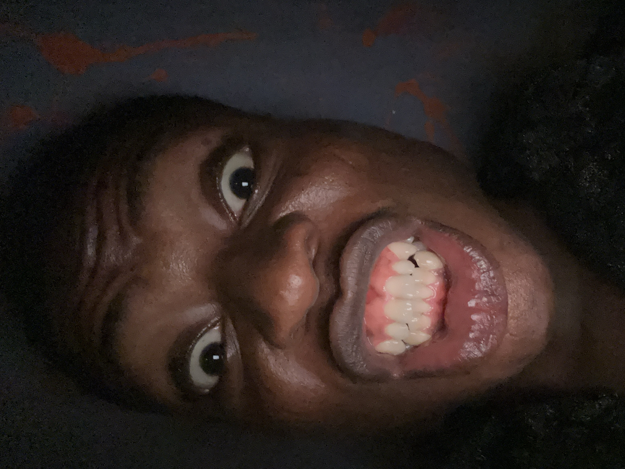 A close-up, tilted photograph depicts a dark-skinned person with their face contorted in a grimace, teeth bared. The person's eyes are wide, and wrinkles are evident on their forehead. The background is dark and blurry, with indistinct red splashes visible at the top. The location is Unknown, Unknown.