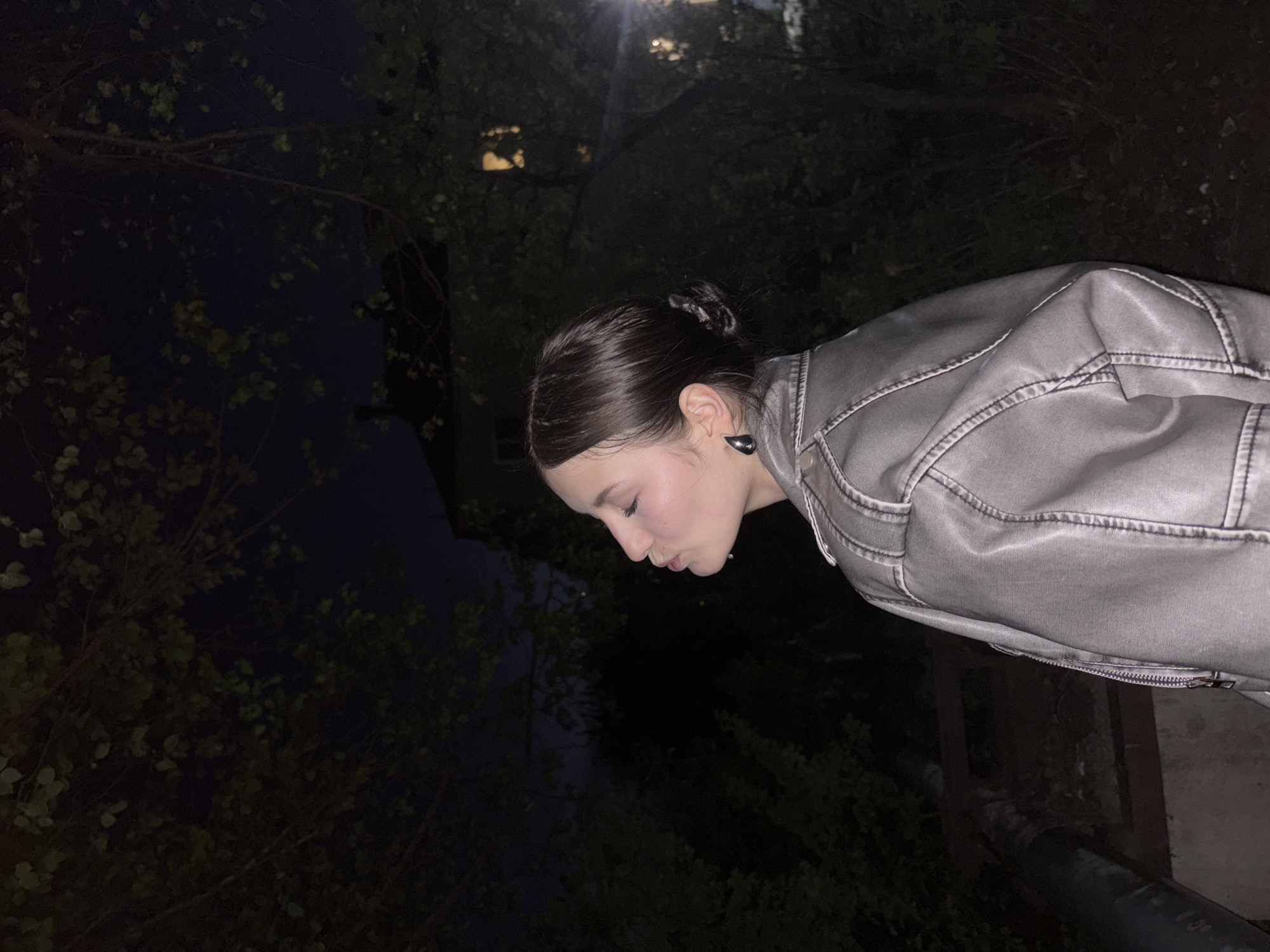 A photograph taken in Фоки, Russia, depicts a young woman outdoors at night. The lighting is dim, possibly illuminated by a distant light source, casting shadows across the scene. She is wearing a light-colored, possibly gray or silver, jacket made of a material that appears to be leather or a leather-like substance. Her hair is neatly arranged in a bun or updo. She is positioned with her head tilted downward and her lips pursed slightly as if in a pensive or kissing gesture.
The background contains foliage, suggesting a park or garden setting. Dark trees surround her on the left and in the background. To her right, a dark shape, possibly a pipe or railing, is visible. The details of the city are not explicitly visible in the frame, but the foliage and the presence of a possible urban element (the pipe) suggest a combination of nature and urban development in Фоки.