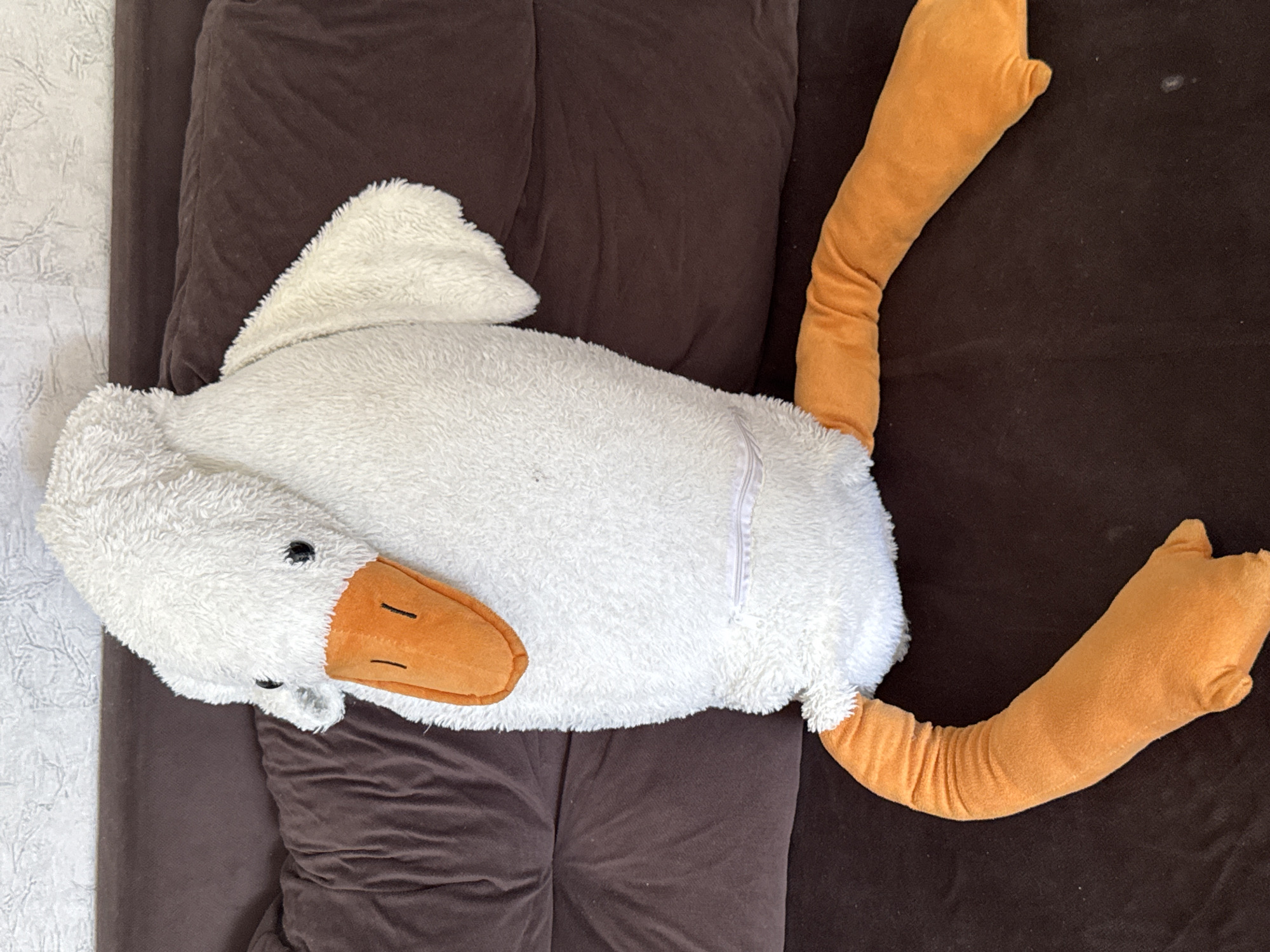 A white plush goose with orange feet and beak is sitting on a brown couch. The goose is facing left, with its head tilted to the left. The goose's legs are extended to the left and right. The couch is in a room with a white, textured wall in the background. The location is Яромінскі сельскі Савет, Belarus.