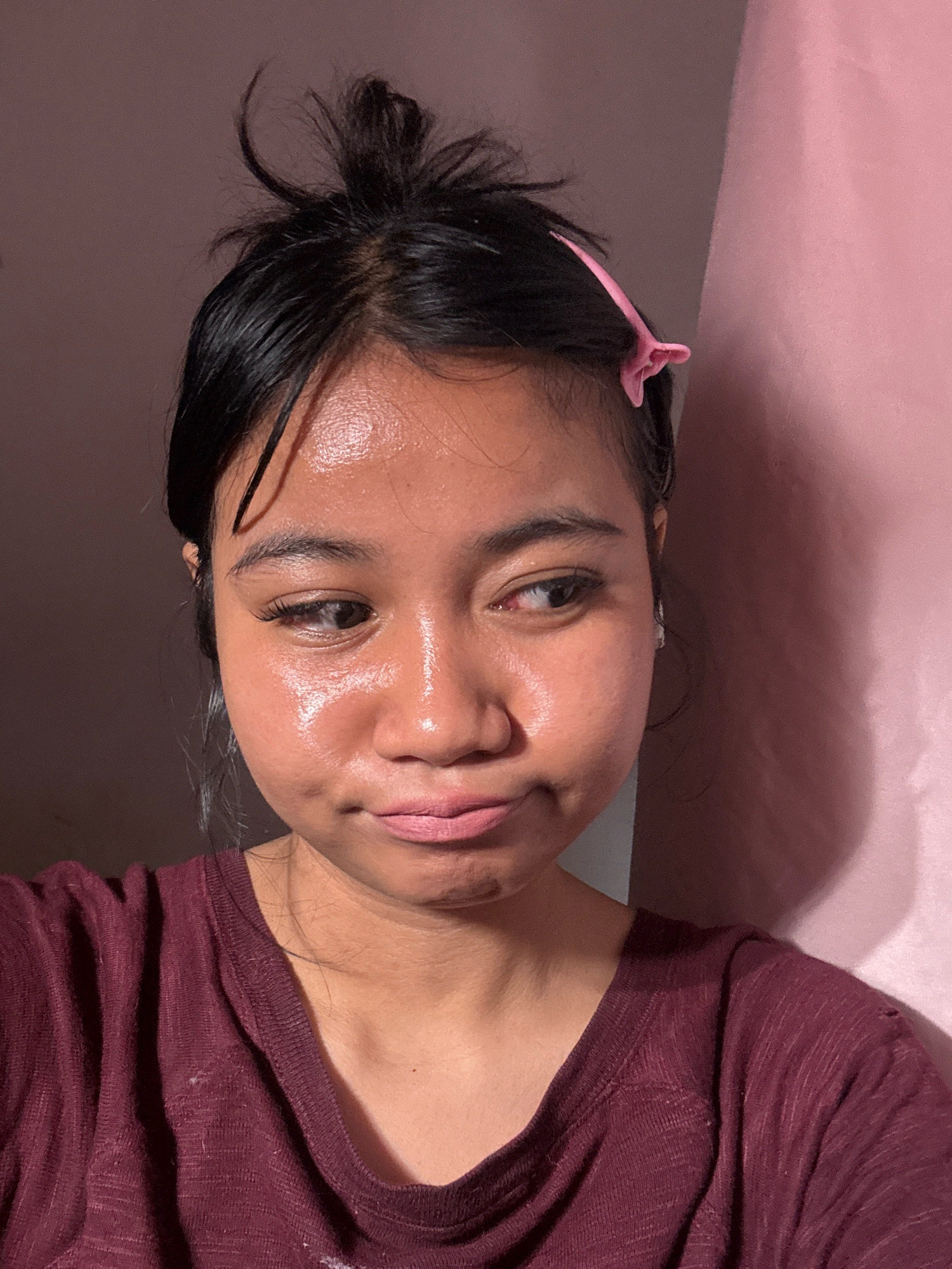A young woman with dark hair pulled up in a messy bun, adorned with a pink hair clip, looks to her right with a slightly pouty expression. Her face is highlighted by a strong light source from the left, emphasizing the sheen on her skin and her eyelashes. She is wearing a dark maroon t-shirt. The background consists of a pink curtain to her right and a darker wall to her left. This image was taken in North Jakarta, Indonesia.