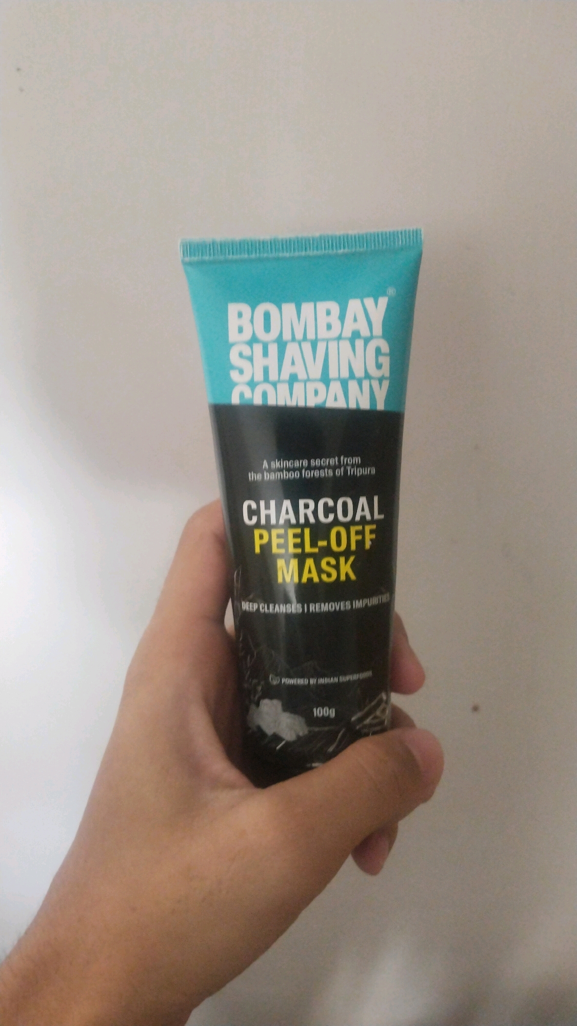 A person's hand holds a tube of Bombay Shaving Company Charcoal Peel-Off Mask against a white wall. The tube is primarily turquoise at the top, with "BOMBAY SHAVING COMPANY" printed in white capital letters. The rest of the tube is black, featuring text indicating it's a "skincare secret from the bamboo forests of Tripura," and "CHARCOAL PEEL-OFF MASK" in white and yellow. Below that, smaller text reads "DEEP CLEANSES | REMOVES IMPURITIES." The tube also shows a "POWERED BY INDIAN SUPERFOODS" logo and "100g" printed on it. The background appears to be a plain, textured white wall, suggesting an indoor setting. The image's composition and the wall's texture are consistent with an environment found in Bihar Sharif, India, a city known for its historical significance and developing urban infrastructure where such personal care products are commonly marketed and used.