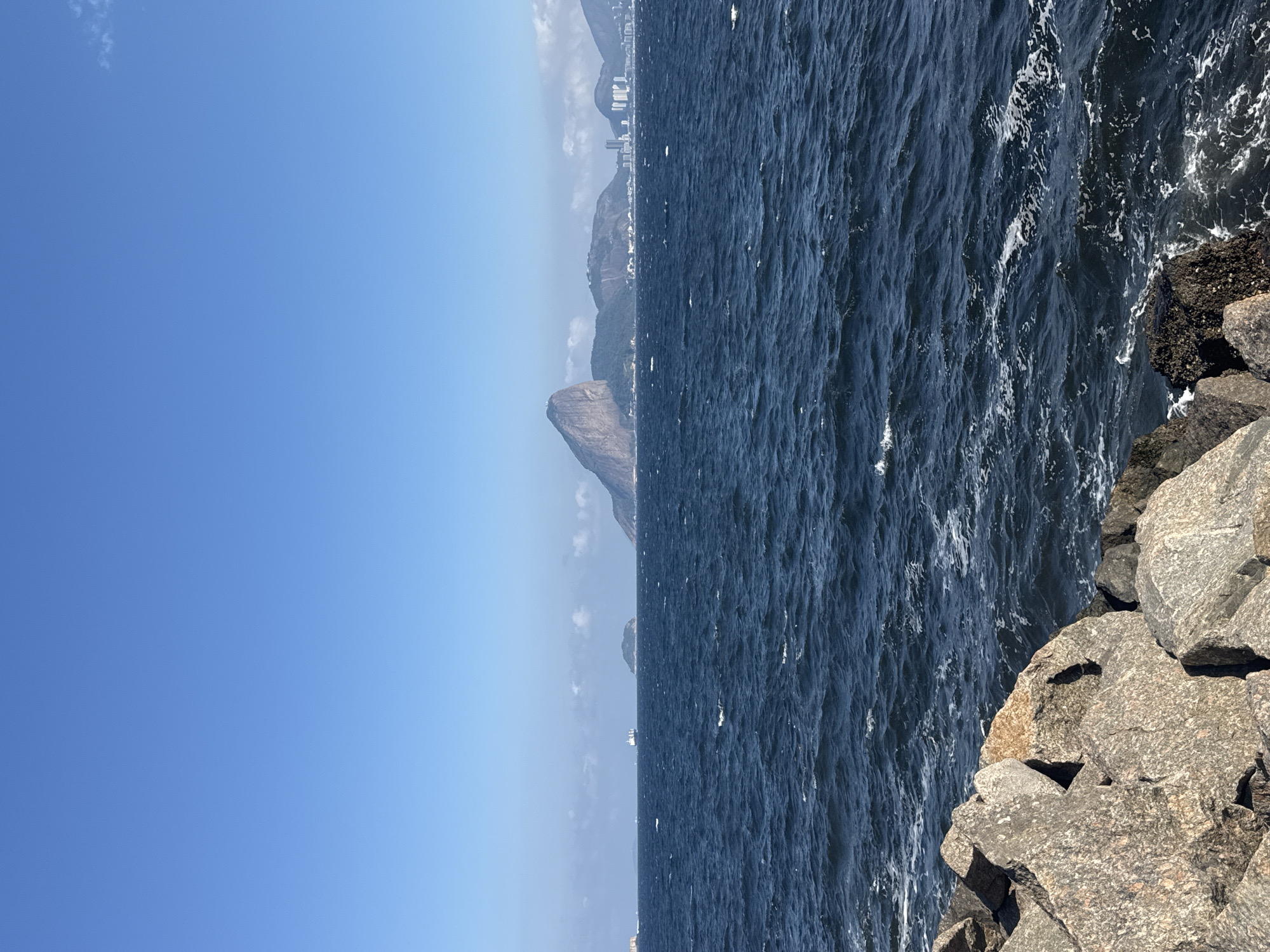 The image shows a panoramic view of Guanabara Bay in Niterói, Brazil. The foreground is dominated by a rocky shoreline, with large, grey stones in sharp focus. The mid-ground consists of the dark blue, rippling water of the bay, with whitecaps indicating a moderate breeze. In the distance, the iconic Sugarloaf Mountain (Pão de Açúcar) is visible on the right, along with other smaller islands and the Niterói skyline with its cluster of buildings. A large cargo ship is positioned on the horizon between Niterói and a smaller, tree-covered island. The sky is a clear, vibrant blue with a few wispy clouds. The scene appears to be captured on a sunny day, likely during daylight hours. No people or specific actions are depicted in the image.