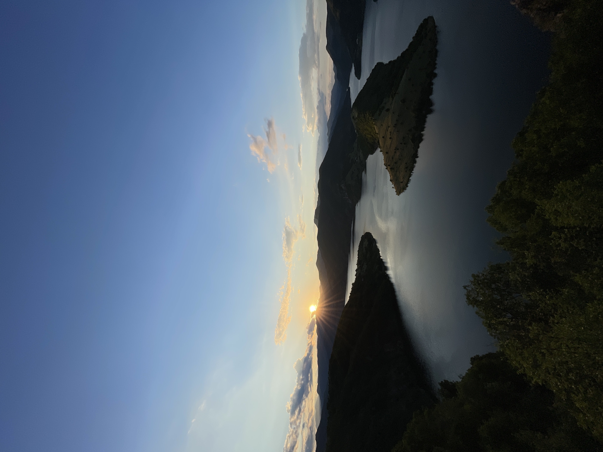 An aerial, wide-angle shot captures a serene sunset over a picturesque landscape in Borovitsa, Bulgaria. The sun, a radiant orb with visible rays, dips below a range of dark, silhouetted mountains in the background, casting a warm glow across the sky and its scattered clouds. A vast body of water, likely a lake or reservoir, dominates the scene, its surface reflecting the soft light of the setting sun. The water forms a winding path through the mountainous terrain, with a prominent, vegetated island in the foreground. The foreground itself is characterized by dense, dark green foliage, suggesting trees and undergrowth, which frames the lower part of the image. The overall atmosphere is tranquil and natural, emphasizing the beauty of the Bulgarian landscape during twilight.