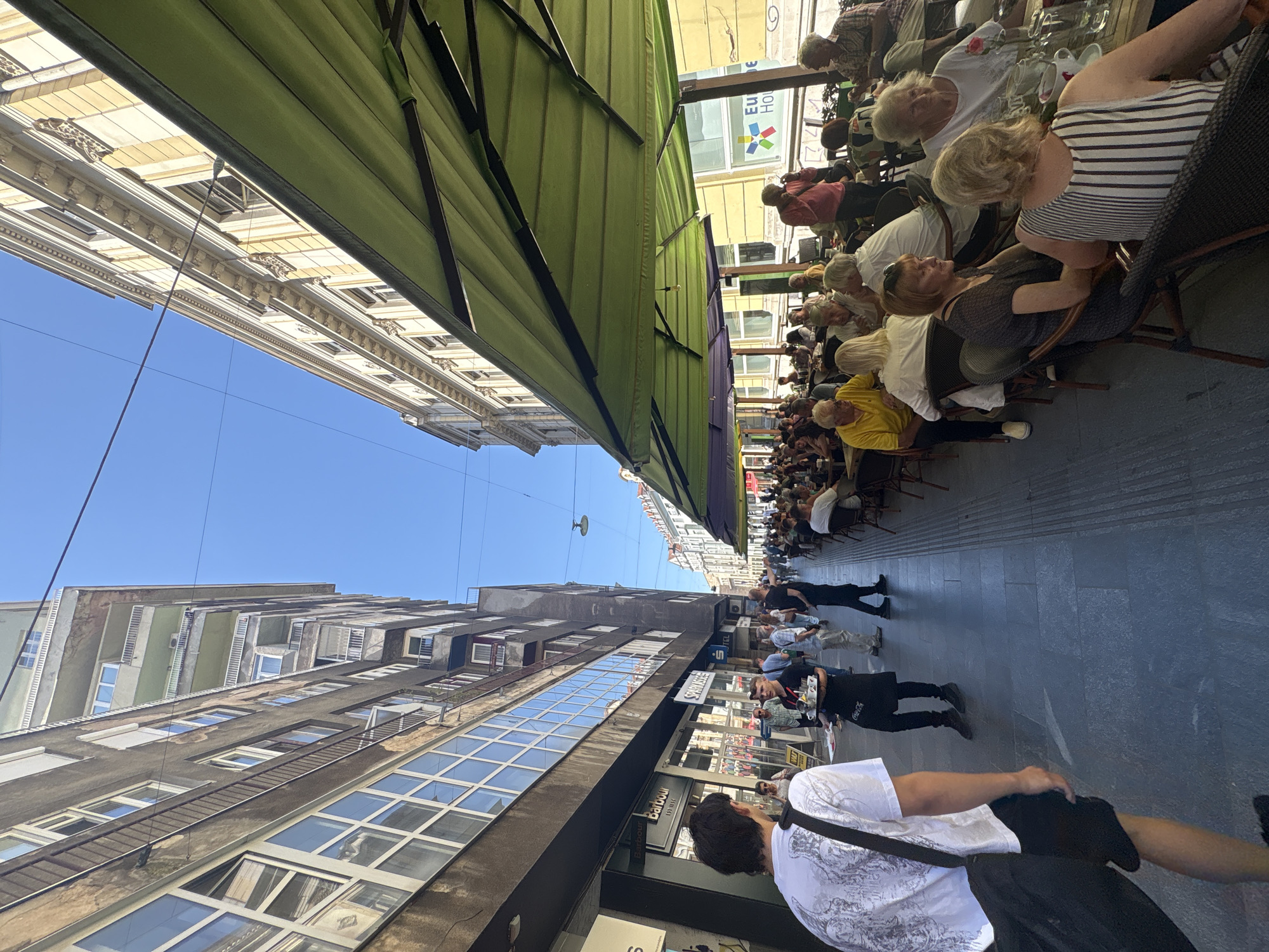 This image captures a lively street scene in Sarajevo, Bosnia and Herzegovina, likely on a sunny day. The perspective is from a person walking down a wide pedestrianized street, looking forward. On the left, a multi-story building with a dark facade and numerous large windows lines the street. Signs for "Barbour" and "Sparkasse" are visible on the ground floor.
To the right, a row of tables and chairs are set up outside a cafe or restaurant, shaded by large green umbrellas. Many people are seated at these tables, engaged in conversation and enjoying drinks and food. A waiter is seen carrying a tray with coffees. The architecture on the right side of the street appears older, with ornate yellow facades and classical detailing.
The sky is clear blue, suggesting fair weather. The street itself is paved with large, dark stones. The overall atmosphere appears relaxed and social, with people enjoying an outdoor dining experience. The scene depicts a typical urban environment with a blend of modern and historic architecture.