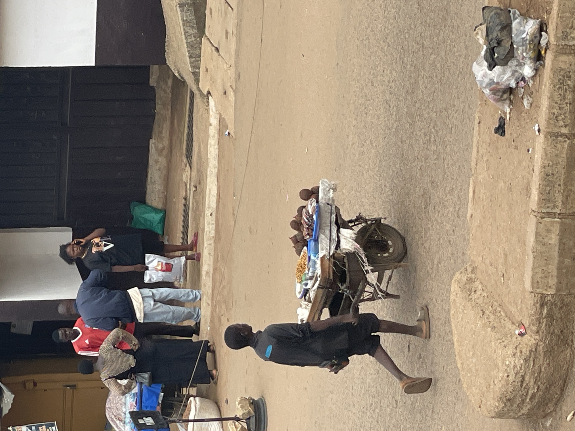The image depicts an outdoor street scene in the city of Unknown, Unknown, under daylight conditions. Five individuals are visible, along with a vendor's cart and architectural elements.
In the foreground, slightly left of center, a young male with dark skin walks rightward, pushing a two-wheeled wooden cart. He wears a dark long-sleeved shirt with a horizontal light blue stripe across the upper back, dark shorts, and light-colored sandals. The cart, covered with a white cloth, carries various food items on plates and in bowls, including yellowish fried snacks and several dark, round items.
In the midground, to the left, a group of four adults stands. A woman with dark skin stands with her back to the camera, wearing a long black dress and a light-colored, textured top, with her hands on her hips. To her right, a man with dark skin wears a red short-sleeved sports jersey with white stripes. Adjacent to him, a second man with dark skin wears a dark blue jacket over a white undershirt and light blue pants, facing away from the camera. To the right of this group, a second woman with dark skin stands, holding a white plastic shopping bag with a red logo in her left hand and a dark mobile phone to her right ear. She wears a black t-shirt with a light graphic and pink flat shoes.
The background features a dark, possibly metallic, building or gate structure on the left, next to a bright green plastic bag on the ground. To its right, a wall transitions from dark brown at its base to white above.
The ground is an unpaved, light brown-tan surface. A metal grate for drainage is visible in the mid-distance. In the lower-left foreground, a low, rounded concrete structure is present. In the lower-right foreground, a concrete curb separates the street from a small area containing a pile of trash in plastic bags and a single dark shoe.