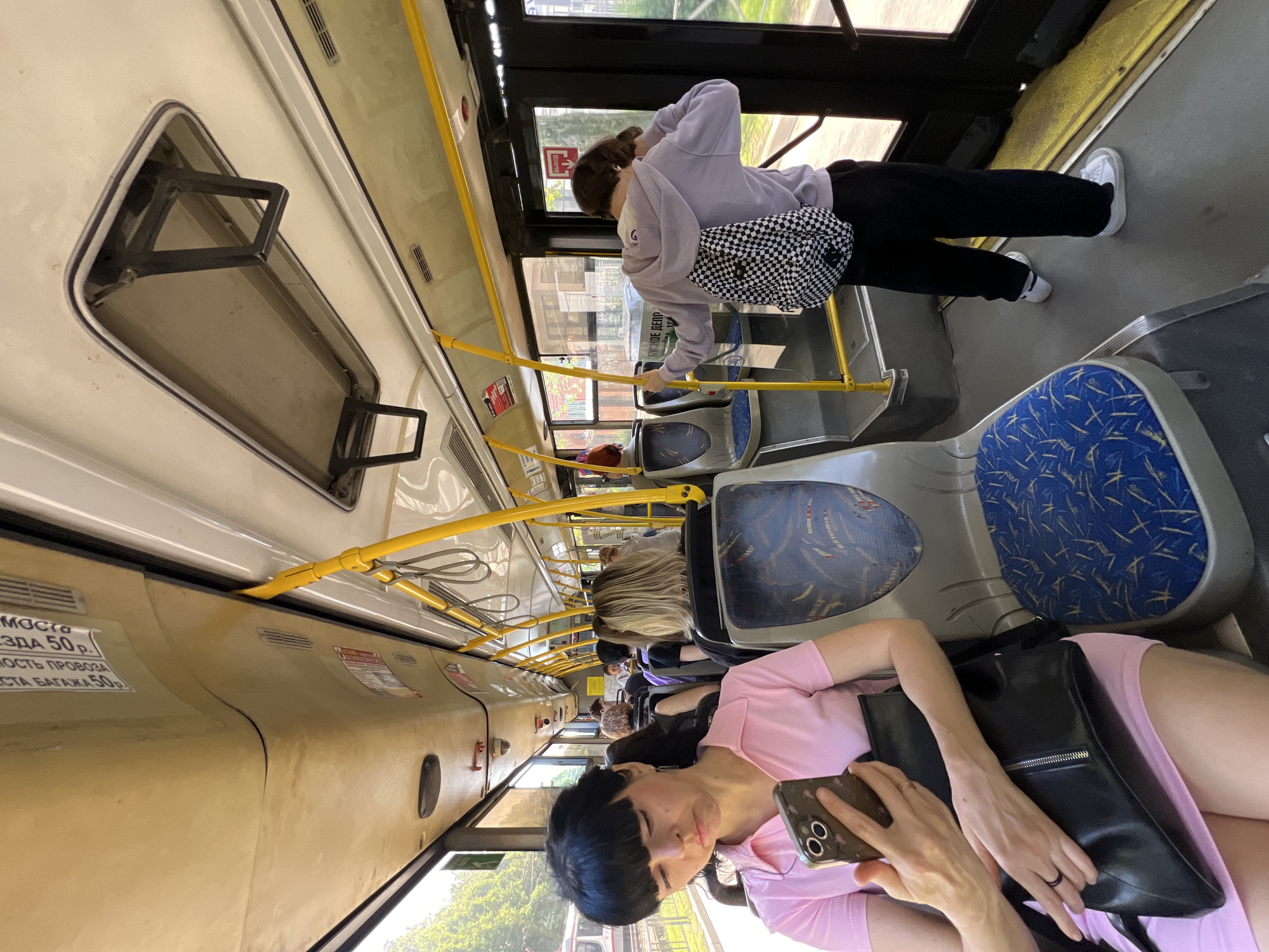The image captures the interior of a bus, likely in the city of Krasnodar, Russia based on the Cyrillic text visible in the upper left corner. The scene includes several individuals: one woman is standing near the bus doors wearing a lavender shirt, black pants, a checkered bag and white sneakers. Another woman is seated towards the front of the bus, wearing a pink dress, holding a smartphone, and with a black purse. There are other passengers visible in the seats behind her. Yellow handrails run the length of the bus, providing support for standing passengers. The seating is upholstered with blue fabric with white patterns. The bus's overhead compartment is visible.