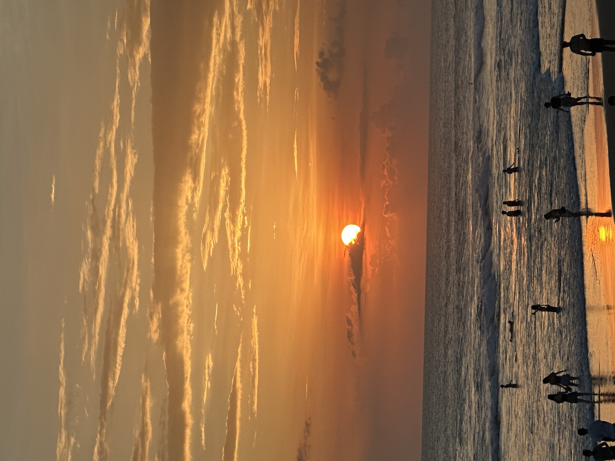 The image depicts a sunset at a beach in Unknown, Unknown. The sun is partially obscured by clouds, casting an orange glow across the sky and reflecting on the water. Silhouetted figures of people are present on the beach and in the water. The beach is sandy. The environment is coastal.