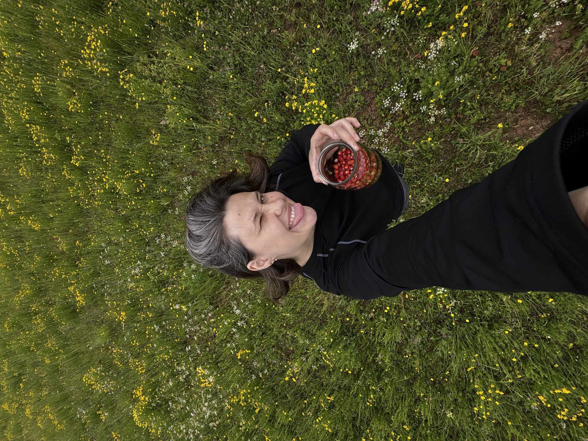 A woman with graying dark hair, wearing a black long-sleeved shirt, is holding a glass jar filled with red berries. She is standing in a grassy field dotted with small yellow and white wildflowers. Her left eye is closed in a wink, and her tongue is sticking out slightly to the right. The camera angle is from above, looking down at the woman.