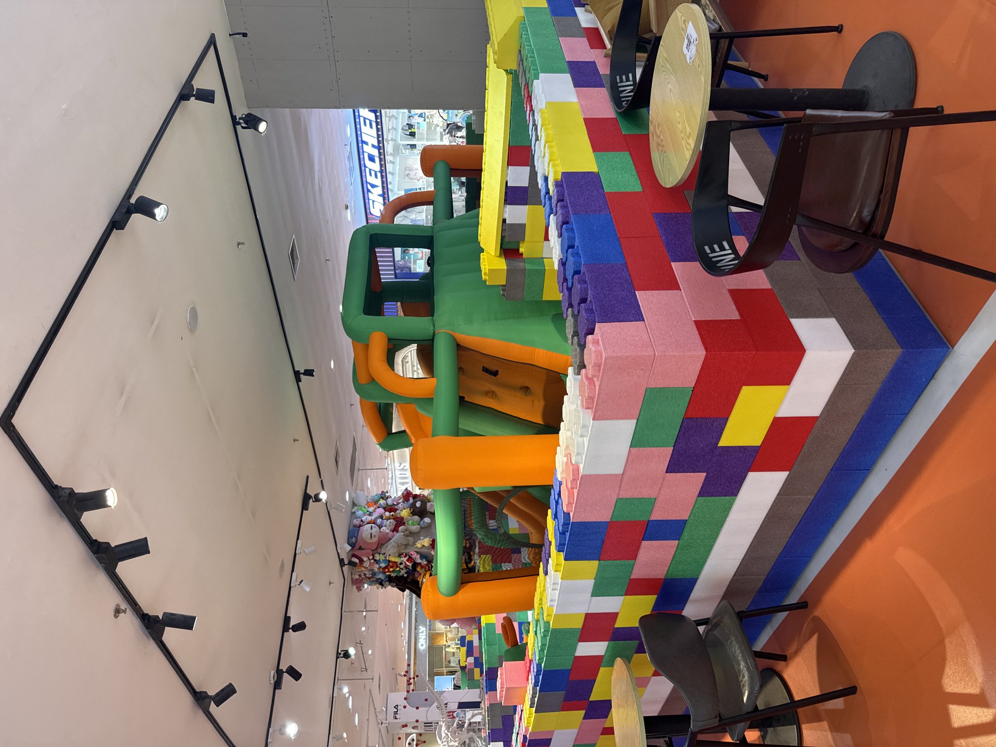 The image depicts an indoor play area with a large, colorful inflatable structure resembling a castle or obstacle course. The structure is primarily green and orange, with various slides and climbing sections. It is surrounded by a low wall constructed from oversized, brightly colored building blocks in shades of pink, red, purple, blue, yellow, green, white, and brown. Two small, round tables with black bases and light-colored wooden tops are visible in the foreground, accompanied by dark-colored chairs, one of which has the word "INE" printed on its backrest. The floor of the play area is a vibrant orange. In the background, store displays are visible, including signage for "SKECHERS" and "ONLY," suggesting the play area is located within a commercial establishment, likely a shopping mall. A tall, dense arrangement of stuffed animals is also visible behind the inflatable structure. The ceiling features a track lighting system with multiple adjustable spotlights. The overall scene suggests a recreational space designed for children within a shopping mall environment in the city of Linghe, China.