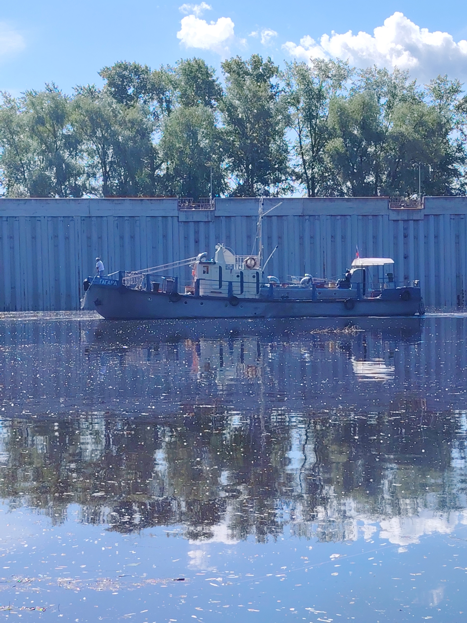 A small, dark-colored boat with a white superstructure is traveling on a body of water. The boat has the name "ГЛАРРА" painted on its side in white letters. A person wearing a white shirt and dark pants is standing on the bow of the boat. The boat is moving in front of a large, grey, corrugated metal wall. Behind the wall, there are numerous green trees. The sky is blue with scattered white clouds. The water's surface is dark and reflective, showing the reflections of the trees, sky, and boat. This scene is likely located in the city of Chaikovsky, Russia, potentially on the Kama River or one of its tributaries, given the characteristic industrial or waterfront infrastructure seen in the image.
