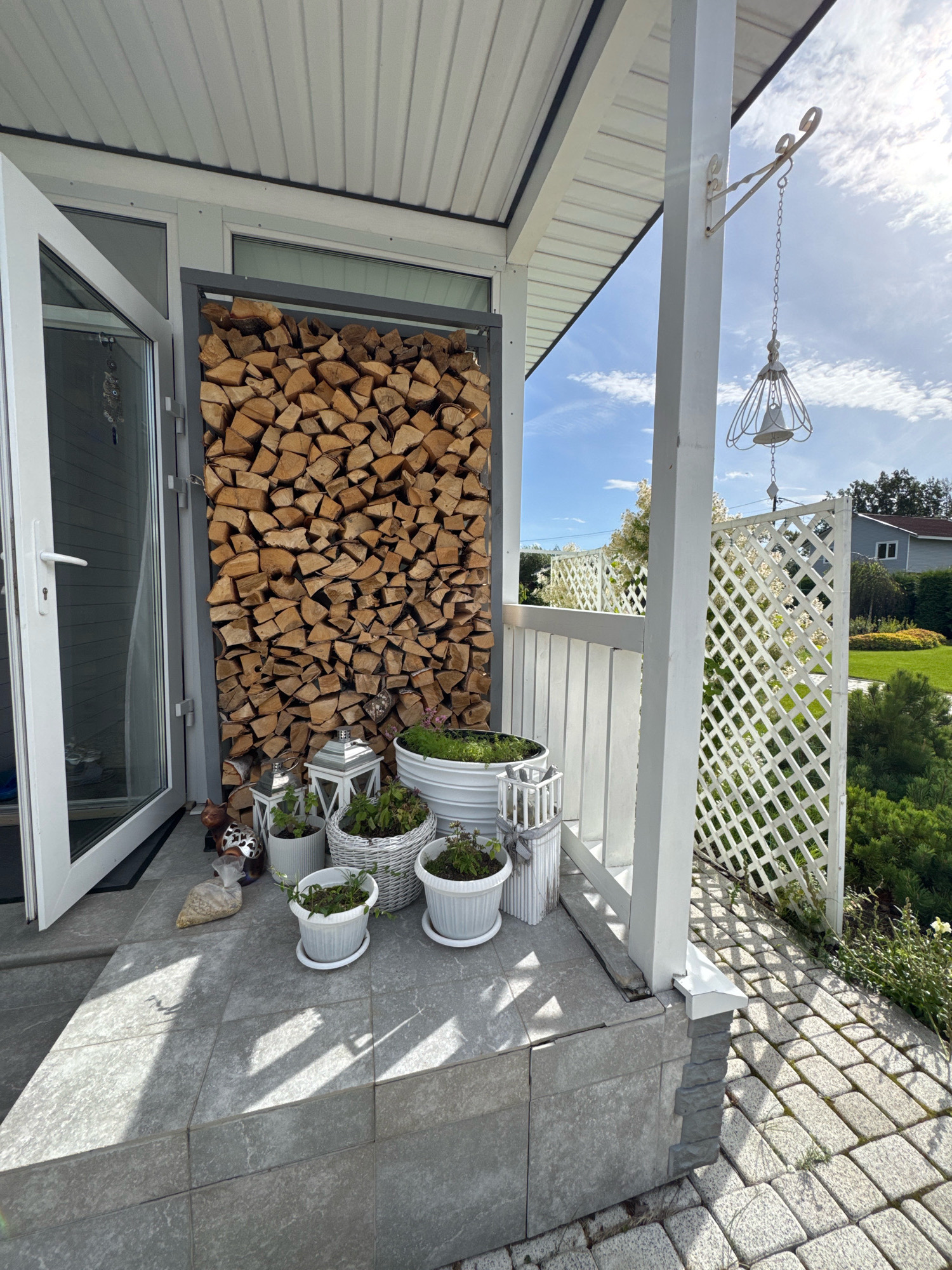 The image shows the exterior of a house in Oliki, Russia. On the porch, there is a large stack of firewood, several white planters with plants, and two white lanterns. A white lattice fence is visible in the background, along with a well-manicured lawn and trees. The sky is blue with some clouds, and the sun is shining, casting shadows on the porch. A white door is open to the left, revealing a glimpse of the interior. A decorative bell hangs from a metal bracket attached to a white porch column. The ground is paved with light gray tiles on the porch and interlocking pavers on the walkway. The overall scene depicts a well-maintained outdoor space, suggesting a pleasant residential area.