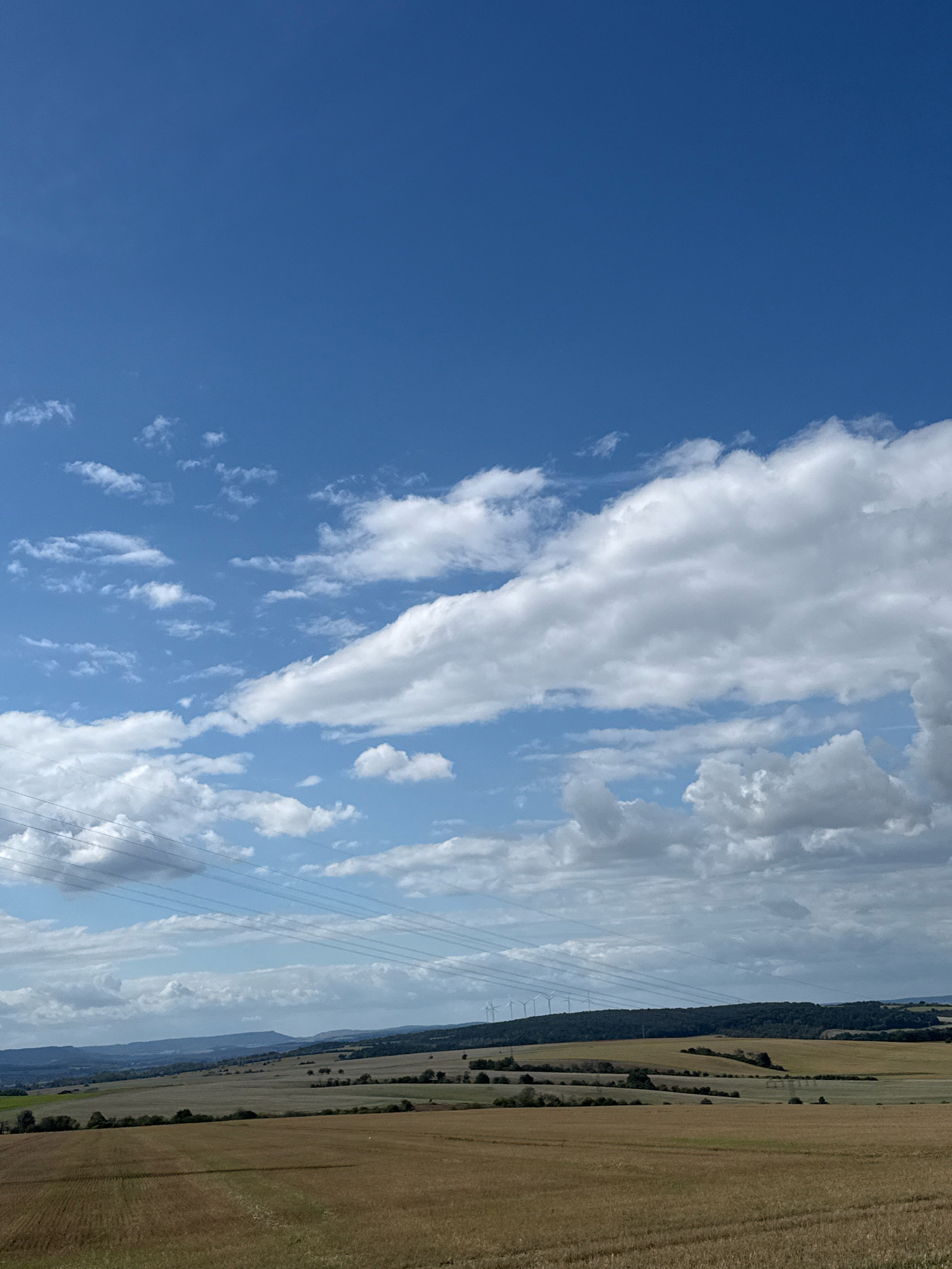 The image depicts a vast, rural landscape under a bright blue sky with scattered white clouds. In the foreground, a field of harvested grain, likely wheat or barley, stretches across the frame, showing the parallel lines left by harvesting machinery. Beyond this field, rolling hills covered in dry grass and scattered trees are visible. A line of power lines cuts across the middle ground, supported by poles. In the distance, a forest borders the horizon, and atop this tree line, several wind turbines are arranged in a row, indicating a wind farm. The scene is set in the vicinity of Kleinfurra, Germany, a location known for its agricultural land and renewable energy installations. The environment is characterized by open fields, gentle topography, and the presence of modern energy infrastructure. There are no people or animals visible in the image.