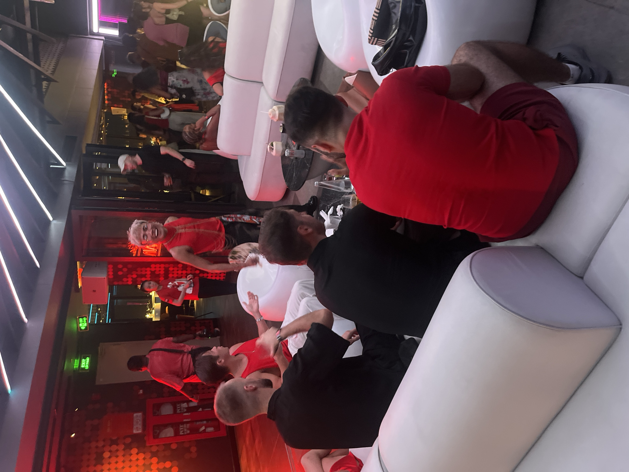 The image depicts a group of people socializing in a dimly lit, modern establishment, possibly a bar or lounge, with predominantly white, modular seating. The scene appears to be indoors, illuminated by artificial lighting, including neon accents and red ambient lighting, suggesting it's nighttime.
Several individuals are visible, many wearing red clothing, which creates a strong visual theme. In the foreground, a person in a red shirt is seated, facing away from the camera. To their left, another person in a black shirt is also seated, their back partially visible. A third individual, also in a black shirt, is seated to the right of the person in red.
Further into the background, a person with blonde hair, wearing a red top, is looking towards the camera with a wide smile and seemingly speaking or laughing. Another person in a red shirt is visible behind them. The overall atmosphere appears lively and social, with people engaged in conversation and enjoying themselves.
The setting includes sleek, contemporary furniture, with white sofas and tables that have dark surfaces. In the background, there are reflections on glass surfaces, indicating windows or mirrored elements. A few drink glasses are visible on the tables. The red lighting casts a warm glow over much of the scene, contributing to a vibrant, possibly celebratory, ambiance. There is no visible weather information as the scene is indoors.
The location context provided is Đà Nẵng, Vietnam, though there are no specific visual cues within the image itself to confirm this location, such as distinct architectural styles or signage in Vietnamese. The presence of "EXIT" signs in English is common in many international locations. The overall impression is one of a modern nightlife venue.