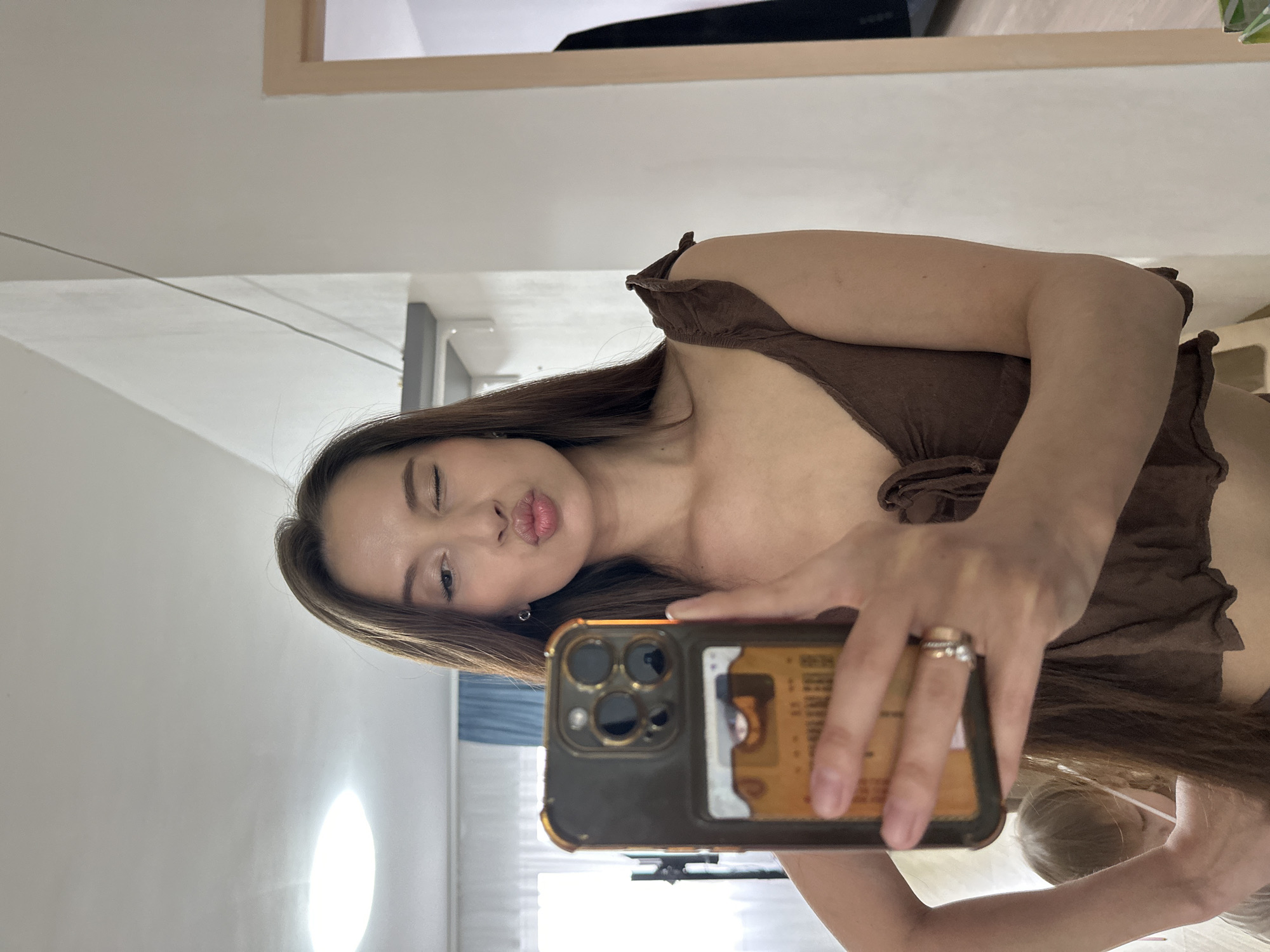 A woman is taking a selfie in a room. She is wearing a brown top and holding her phone, a gold and black iPhone, in her right hand. Her long brown hair is partially covering her face. She is making a kissing face with one eye winked. The background shows a room interior with white walls, a window, and a mirror frame. No specific location information related to Yekaterinburg, Russia is discernible in the image.