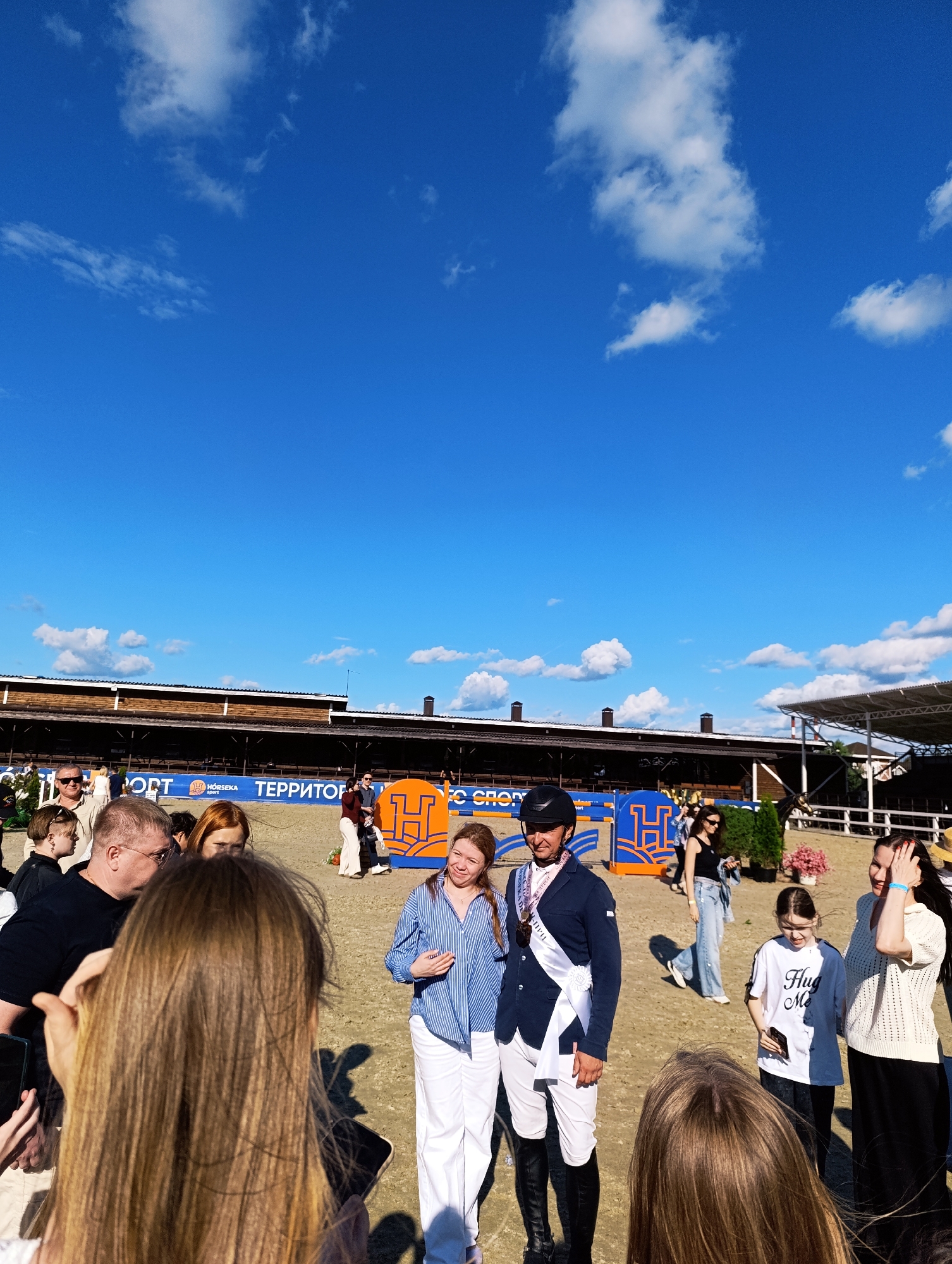This image captures a scene at an equestrian event in Ivanovskoe, Russia, under a bright blue sky with scattered white clouds. In the foreground, several people are gathered, some with their backs to the camera, suggesting they are spectators or participants. A woman in a blue and white striped shirt and white pants stands to the right of a man in a dark blue equestrian jacket and helmet, who is wearing a white sash. This man is likely a competitor or award recipient. Other individuals, including a young girl in a white t-shirt and a woman in a white knitted top, are also present. In the background, there are equestrian obstacles, a grandstand-like structure, and banners displaying text. The ground appears to be sandy, typical of an arena. The overall atmosphere suggests a sunny day at a sporting event.