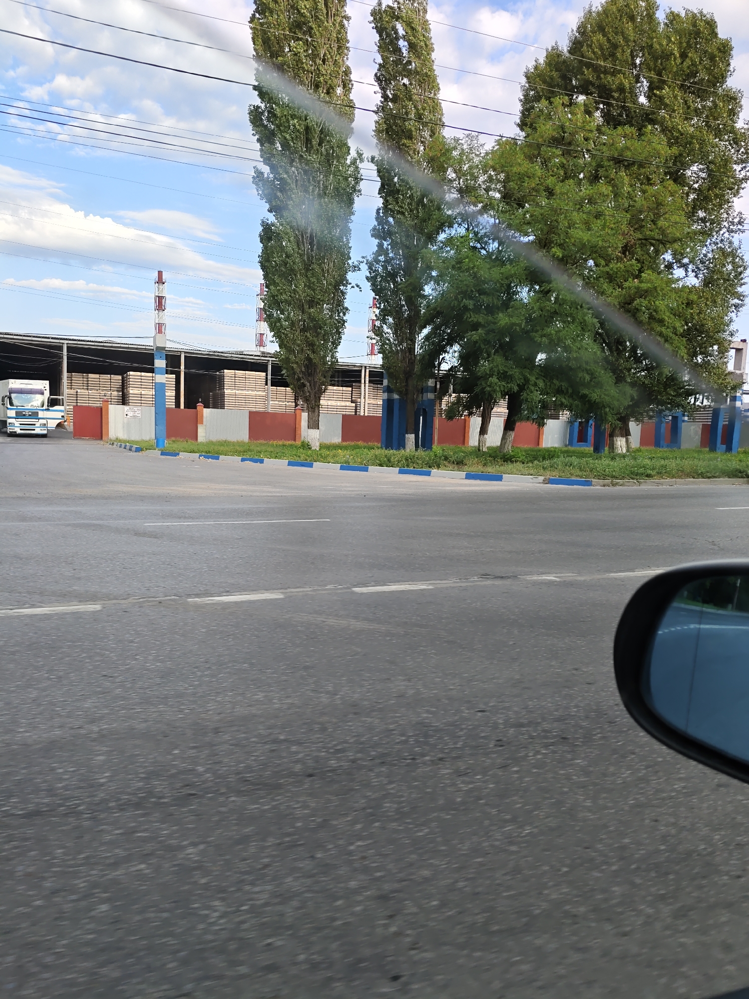 The image depicts a view from a vehicle driving on a paved road in Novocherkassk, Russia. In the background, a large industrial building with a corrugated metal roof is visible. This building is filled with stacks of lumber. Two tall, narrow industrial chimneys with red and white banding rise above the building. Along the front of the building, a fence constructed of red and gray panels separates it from the road. The fence is interspersed with blue and white vertical posts. A white truck with a blue stripe is parked to the left of the building. Tall, slender trees and smaller, bushy trees line the area in front of the industrial building. The road surface is asphalt, with white lane markings. A side-view mirror of the vehicle is visible in the lower right corner of the image, reflecting the sky and some trees. The sky is partly cloudy.