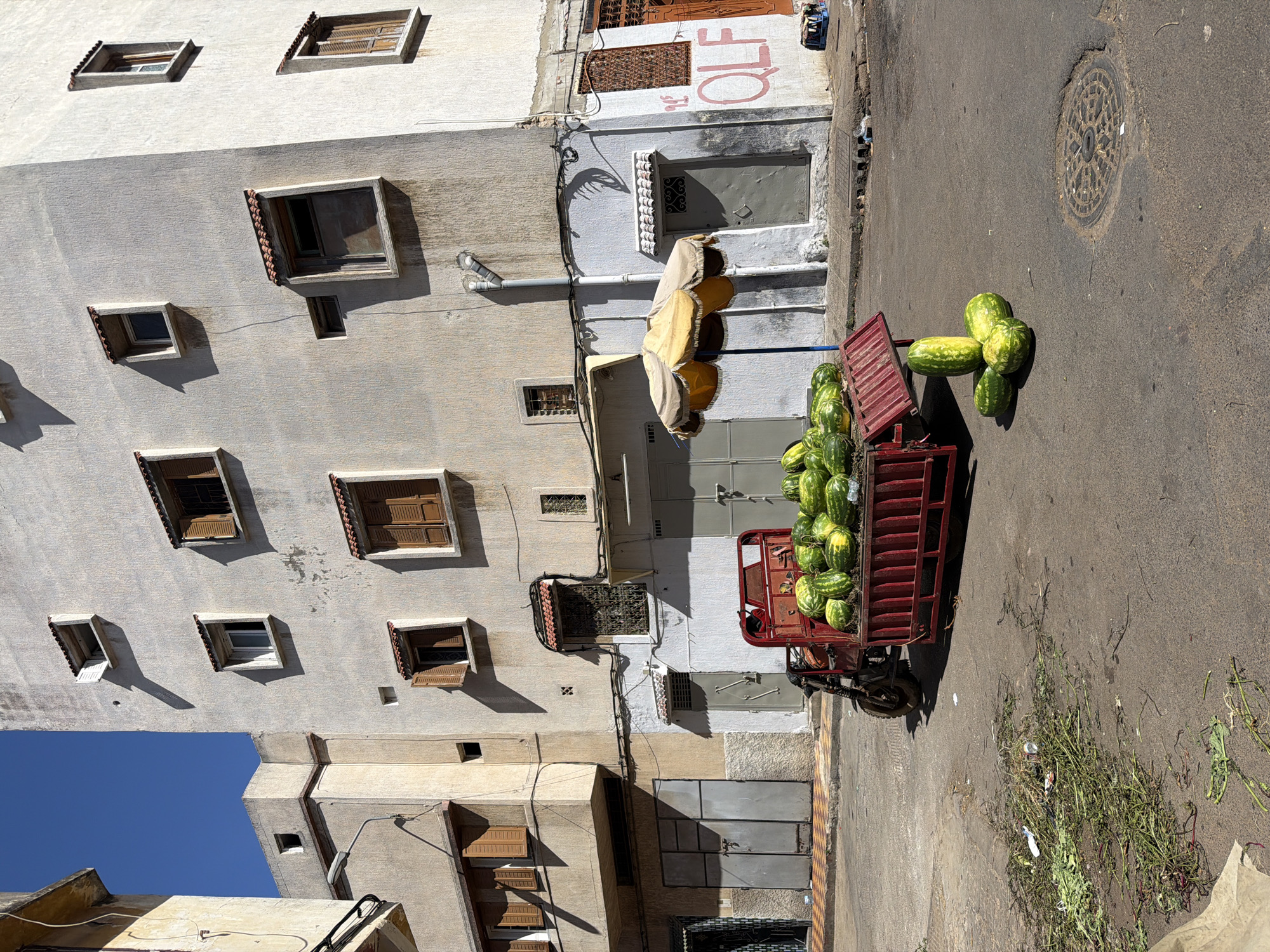 Captured from an elevated perspective, the image presents a street scene in Kenitra, Morocco, showcasing a red motorized cart laden with watermelons, parked near a building facade. The building's wall is white with numerous, varied windows, interspersed with the architectural detailing, casting shadows under a clear blue sky.
A vendor is on the cart. Piles of harvested watermelon stalks and remnants are scattered on the asphalt.
The background contains other residential or commercial buildings, marked with various doors and windows. There is an inscription that says OLF with red painted letters.
The setting suggests a normal day-to-day commercial activity within the urban context of Kenitra, Morocco.