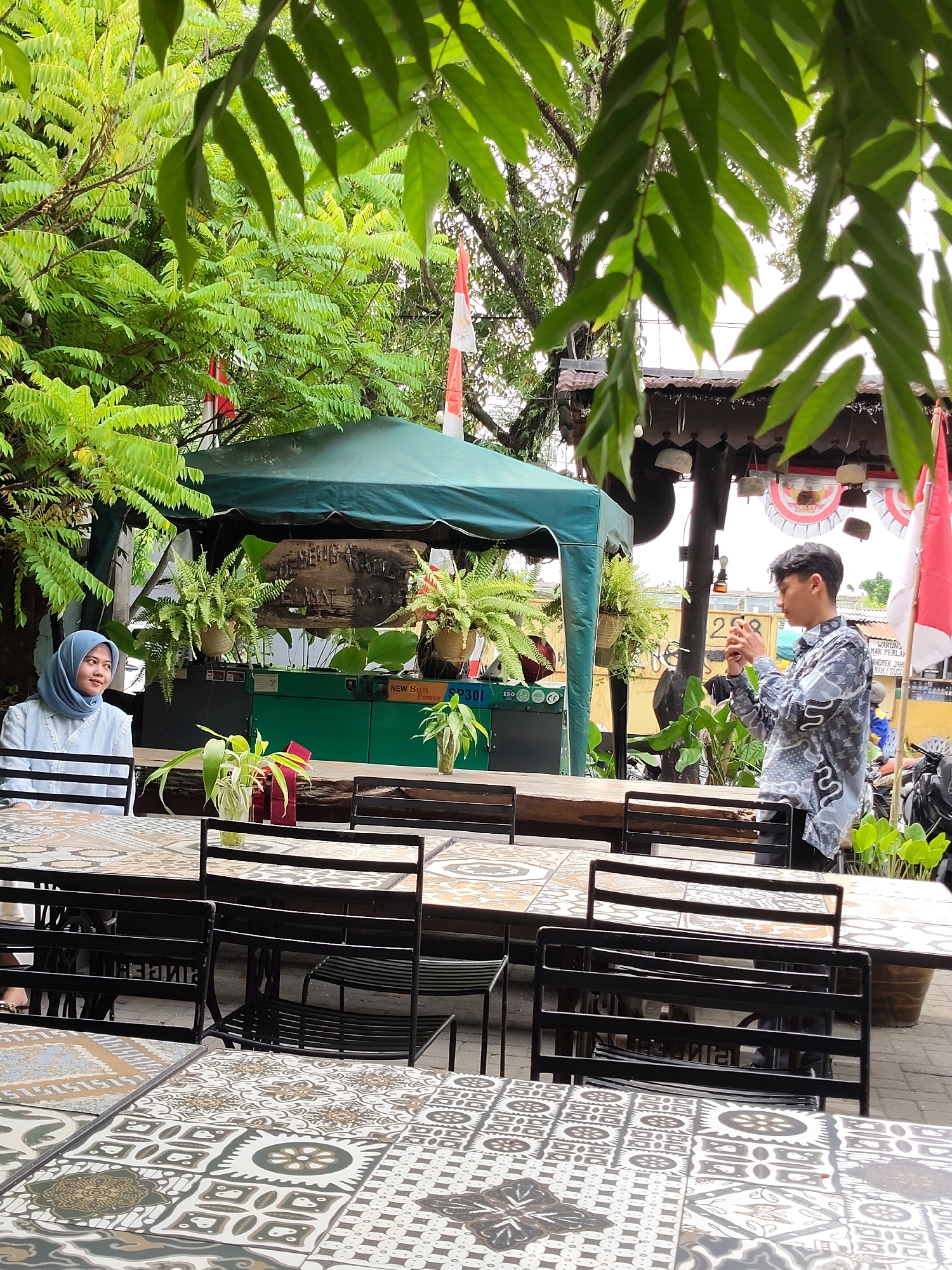 This is a medium shot taken outdoors, possibly at a cafe or restaurant in Medan, Indonesia. In the foreground, several tables are set with black metal chairs. The tabletops have intricate, tiled patterns in shades of beige, brown, and white.
To the left, a young woman wearing a light blue hijab and a matching blouse sits at one of the tables, looking off to the side. In the background, a young man in a patterned shirt is standing, holding a smartphone and appearing to take a picture. He is positioned behind a wooden table.
Behind the tables, a green canopy tent is set up, with potted ferns and other plants visible. A wooden sign with Indonesian text is mounted on the tent structure. In the background, there are also hints of red and white flags, and possibly part of a building with a dark roof. The overall impression is an outdoor dining or resting area, with lush greenery framing the scene. The presence of Indonesian text on the sign and the style of dress suggest a location within Indonesia.