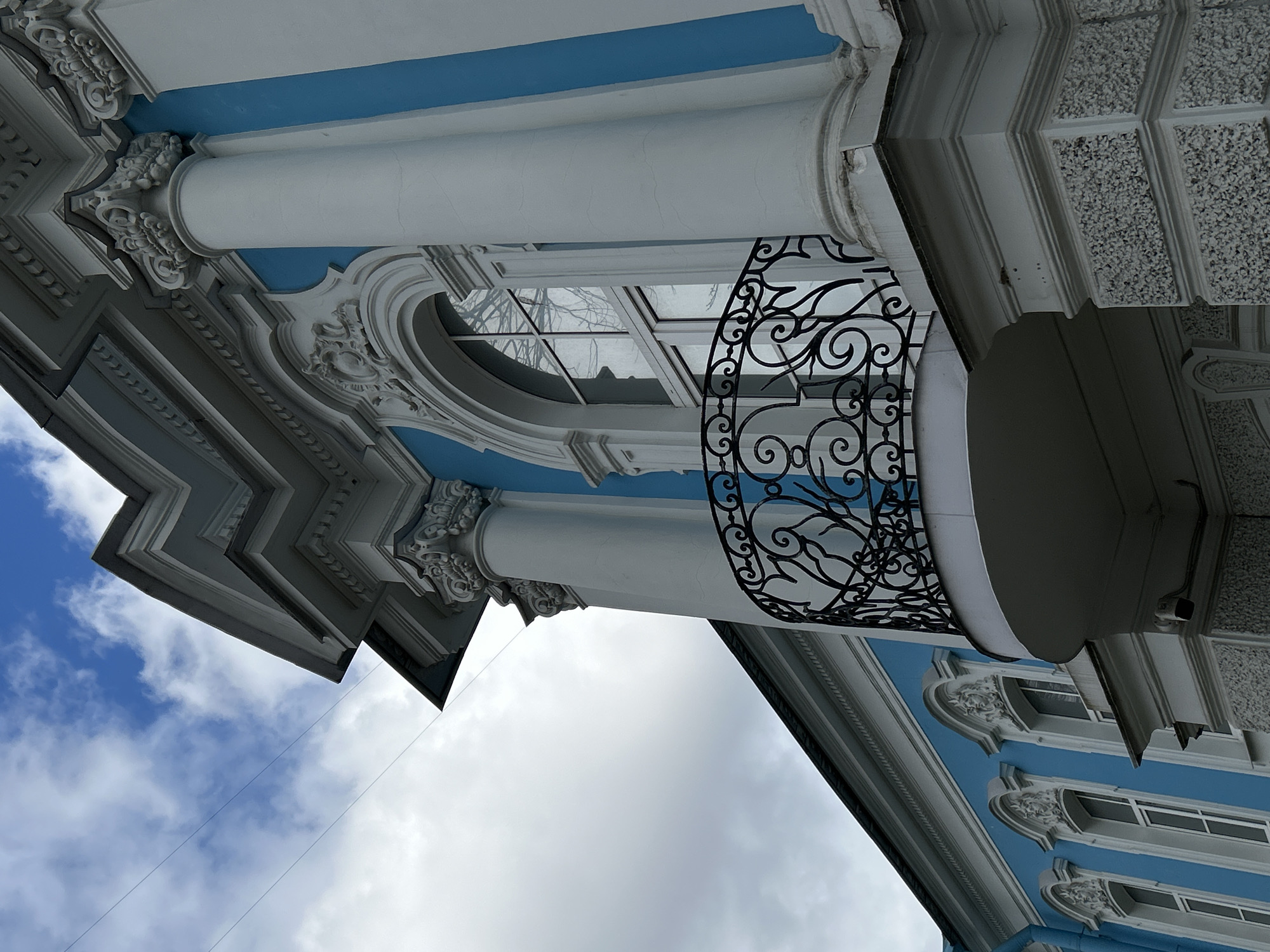 The image shows a low-angle upward view of an ornate building facade in Saint Petersburg, Russia. The building is predominantly blue and white, featuring classical architectural elements like columns, moldings, and decorative relief work. A wrought iron balcony with scrollwork detail is visible. The facade also includes an oval window. The sky is visible in the background, showing both blue sky and clouds. A thin wire is strung across the view towards the left side of the image.