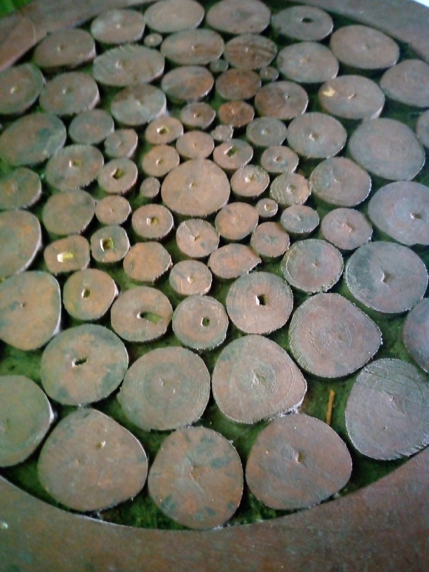 The image shows a close-up of a circular object with a textured surface. The object appears to be made of many small, roughly circular pieces of wood or similar material arranged in a mosaic pattern. These pieces are varying in size and have a natural, rough texture, with some showing visible wood grain and circular growth rings. Many of the wood pieces have a small hole or indentation in their center. The overall color palette is dominated by shades of brown, gray, and green, with the green likely representing a background material. The lighting suggests an indoor or shaded outdoor environment. The image has been taken from a slightly elevated angle looking down onto the surface.
Without additional information or context, it is impossible to determine the specific location of this object within the city of Sumberwono, Indonesia.